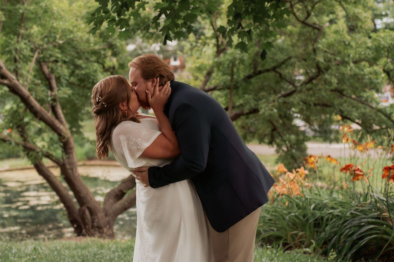 A bride and groom share a romantic kiss outdoors surrounded by lush green trees and vibrant orange daylilies.