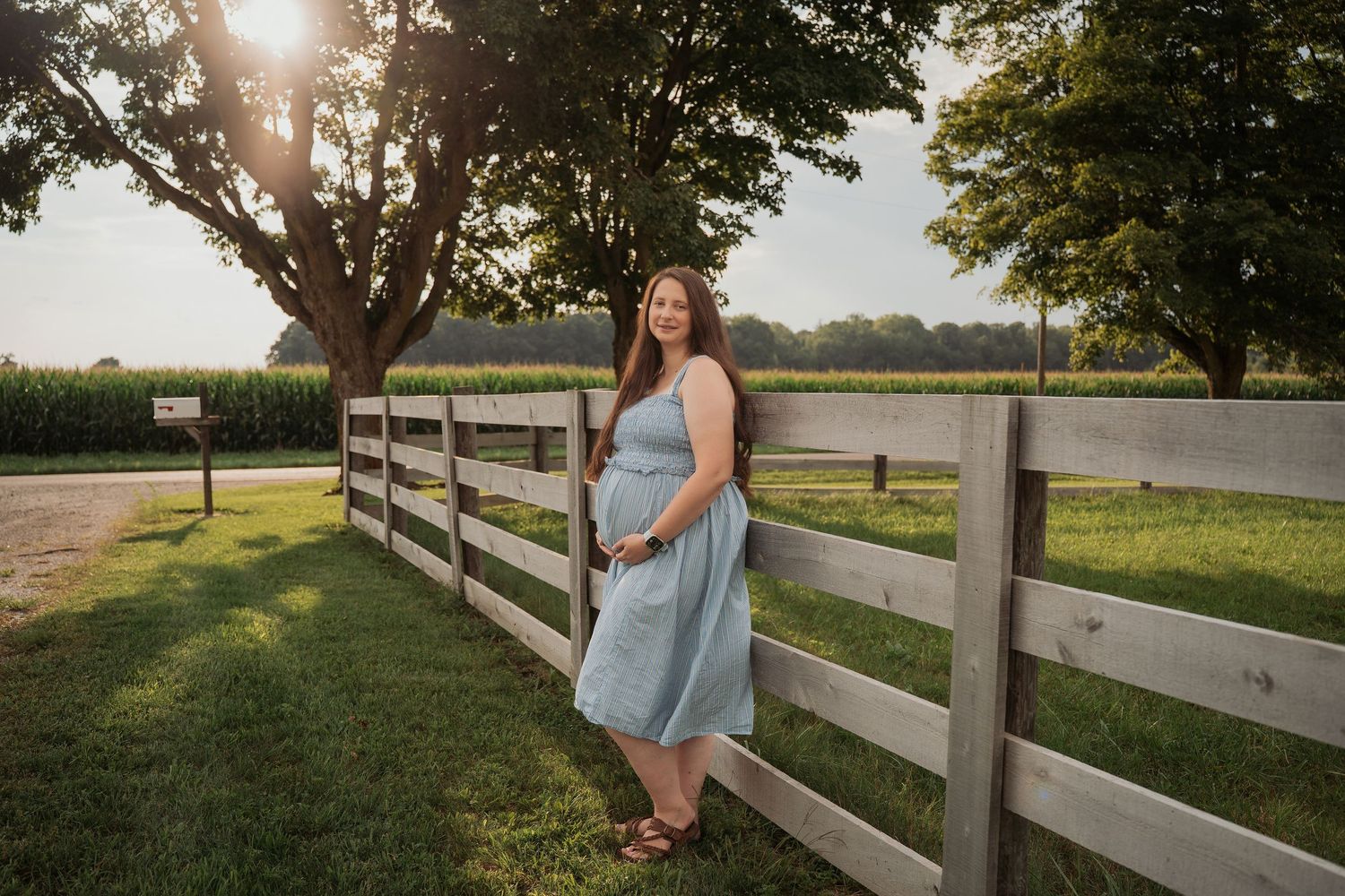 Woman in blue dress leaning on wooden fence at golden hour on a farm with corn fields and trees.