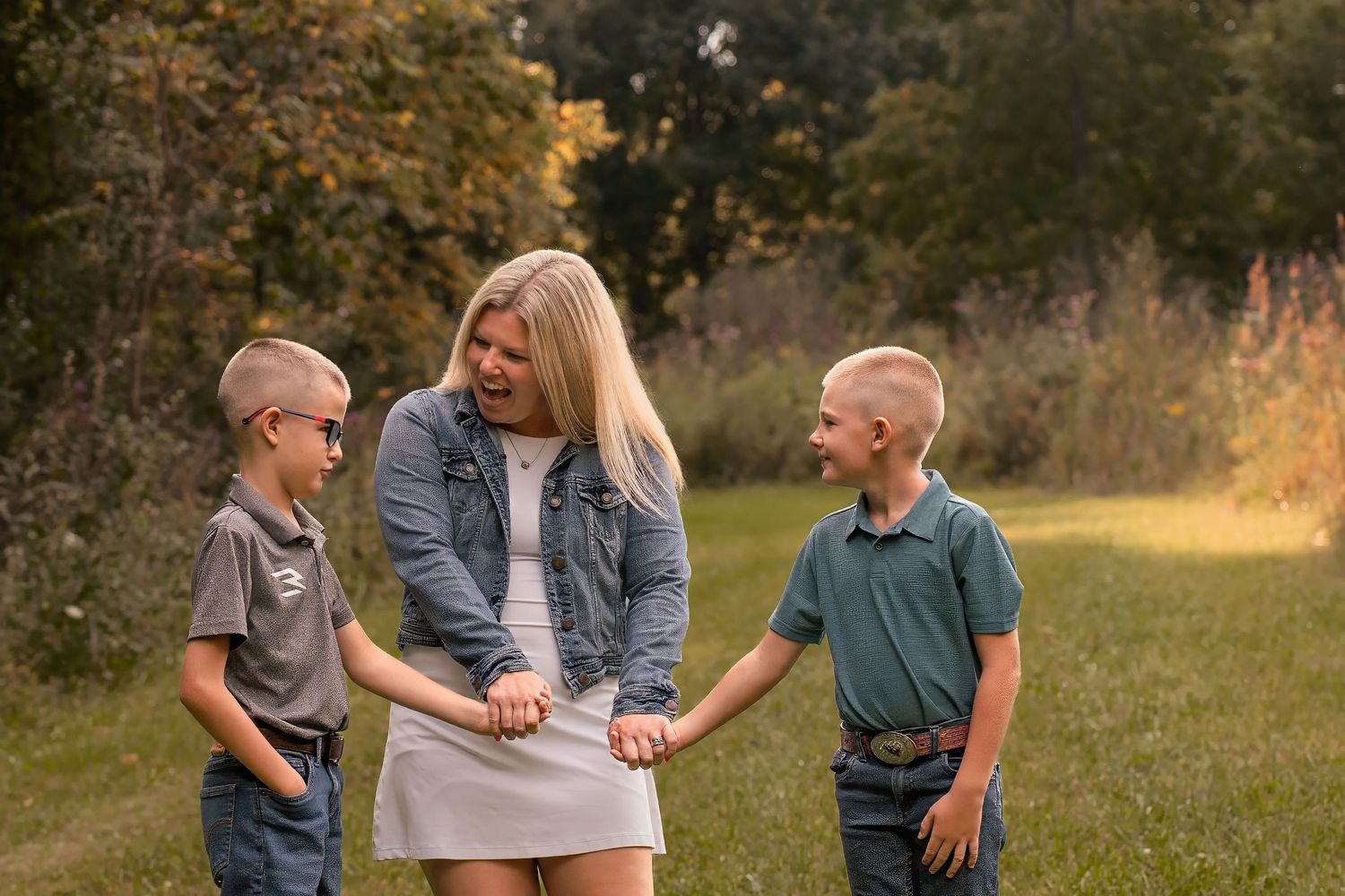 A blonde woman holds hands with two young boys with buzzcuts on a grassy path surrounded by autumn trees.