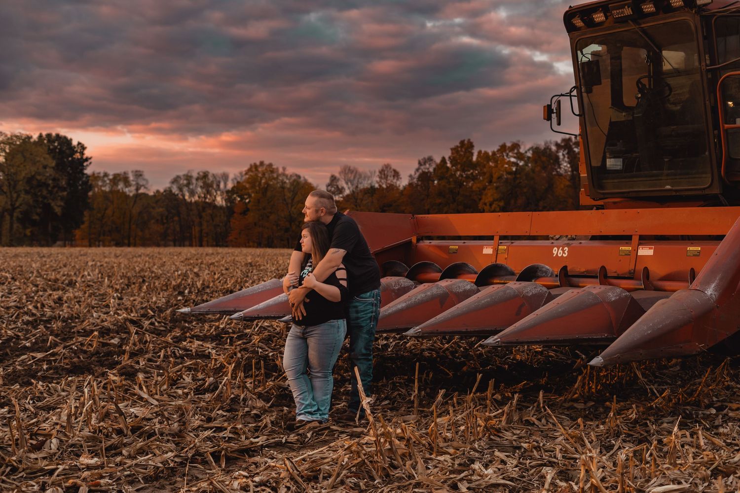 Couple embraces near harvesting equipment during sunset in cornfield.