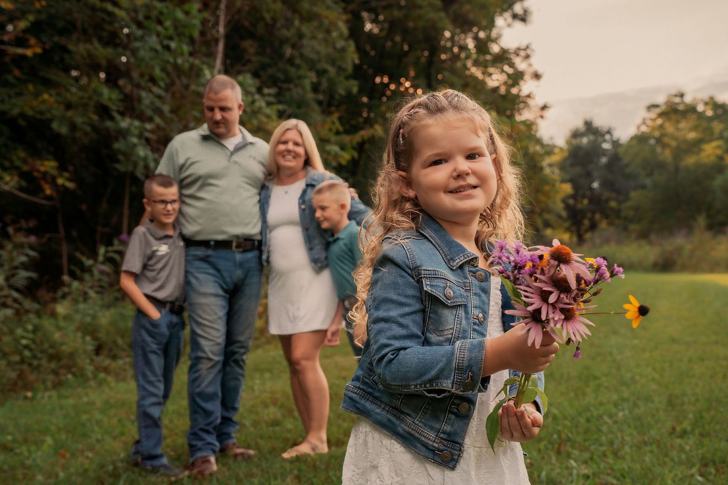 A young girl holds wildflowers while her family smiles behind her in a lush green field during a portrait session.