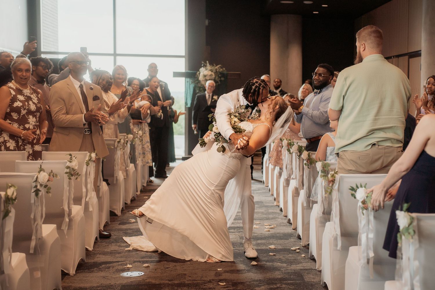 Bride and groom share a joyful moment in the wedding aisle surrounded by clapping guests in a modern venue.
