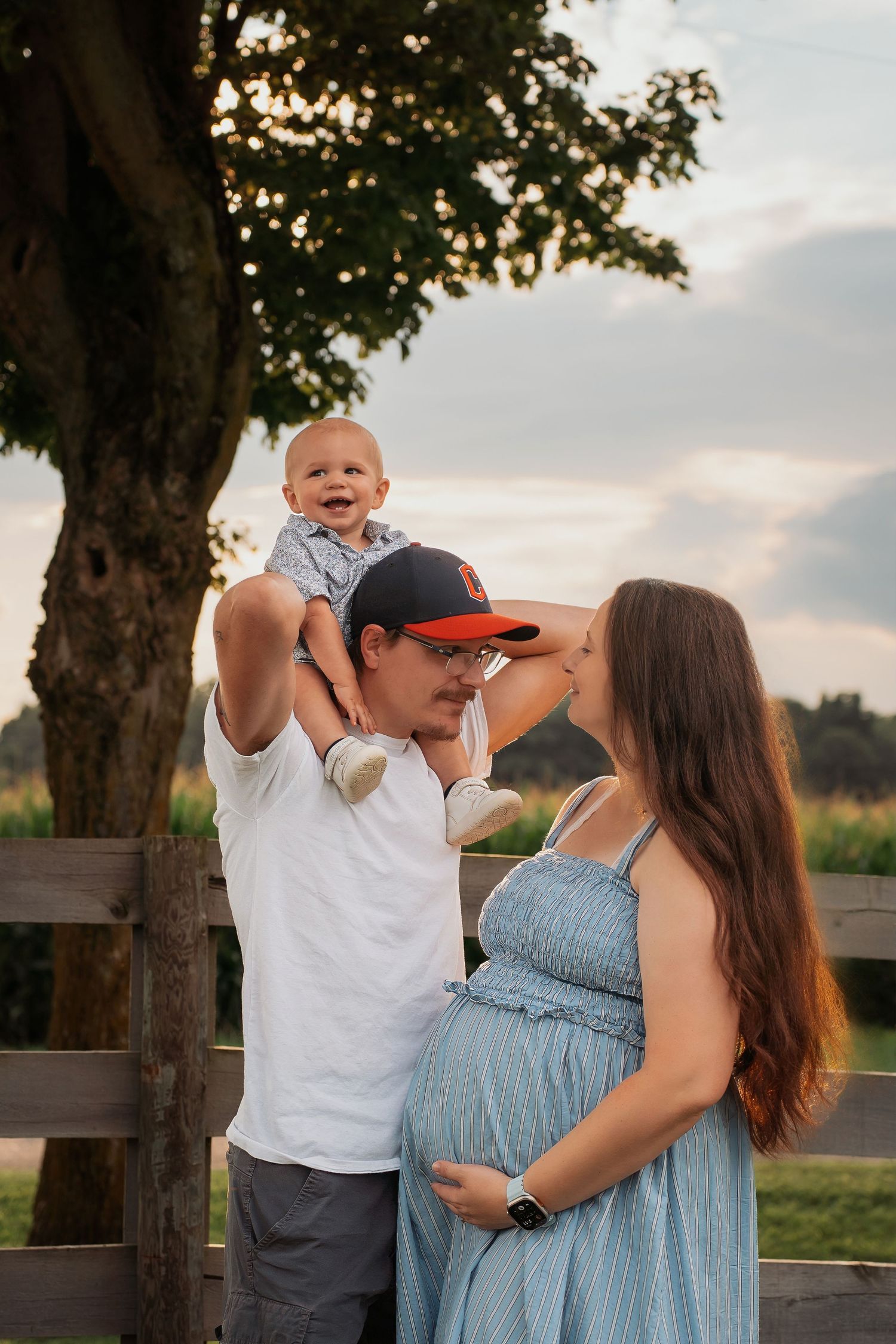 Pregnant woman in blue dress with husband holding baby up outdoors near wooden fence at golden hour.