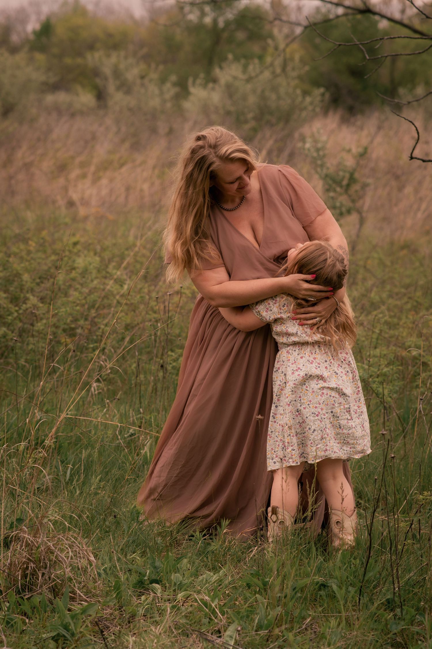 A mother in a mauve dress embraces her young daughter in a floral skirt in a lush green field.