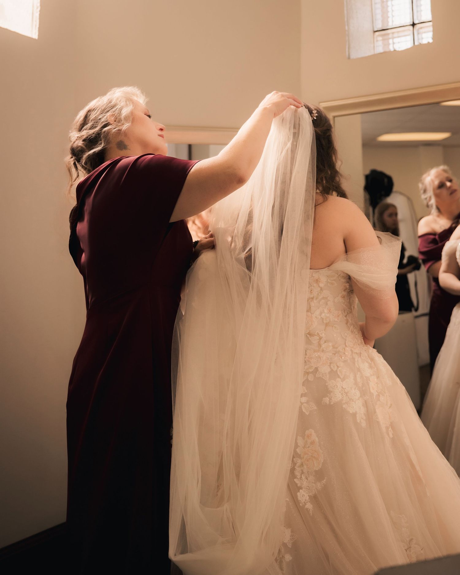 A woman in burgundy dress adjusts a bride's cathedral veil in a bright dressing room before the wedding ceremony.