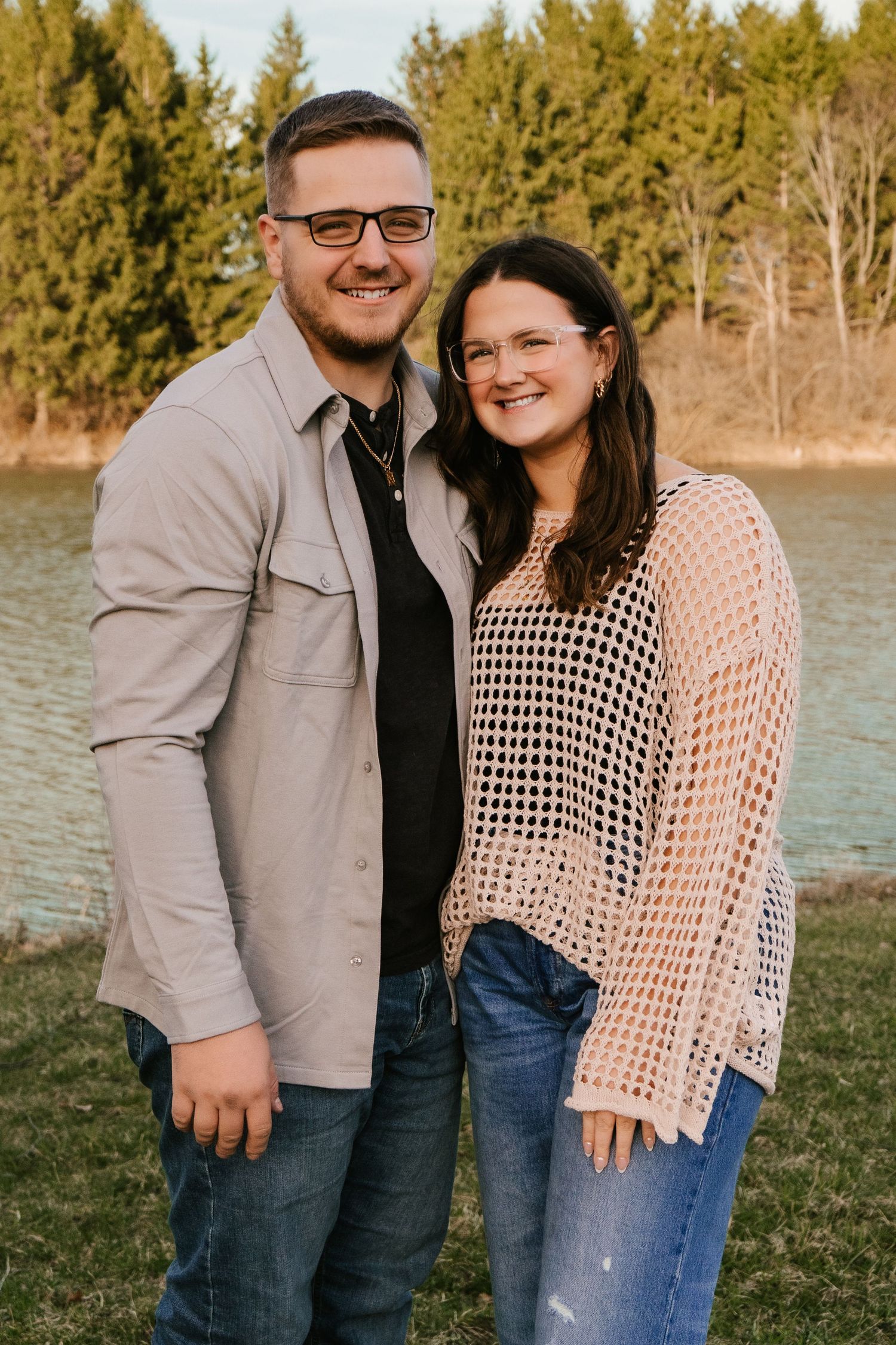 Couple poses by waterfront with evergreen trees in background during golden hour.