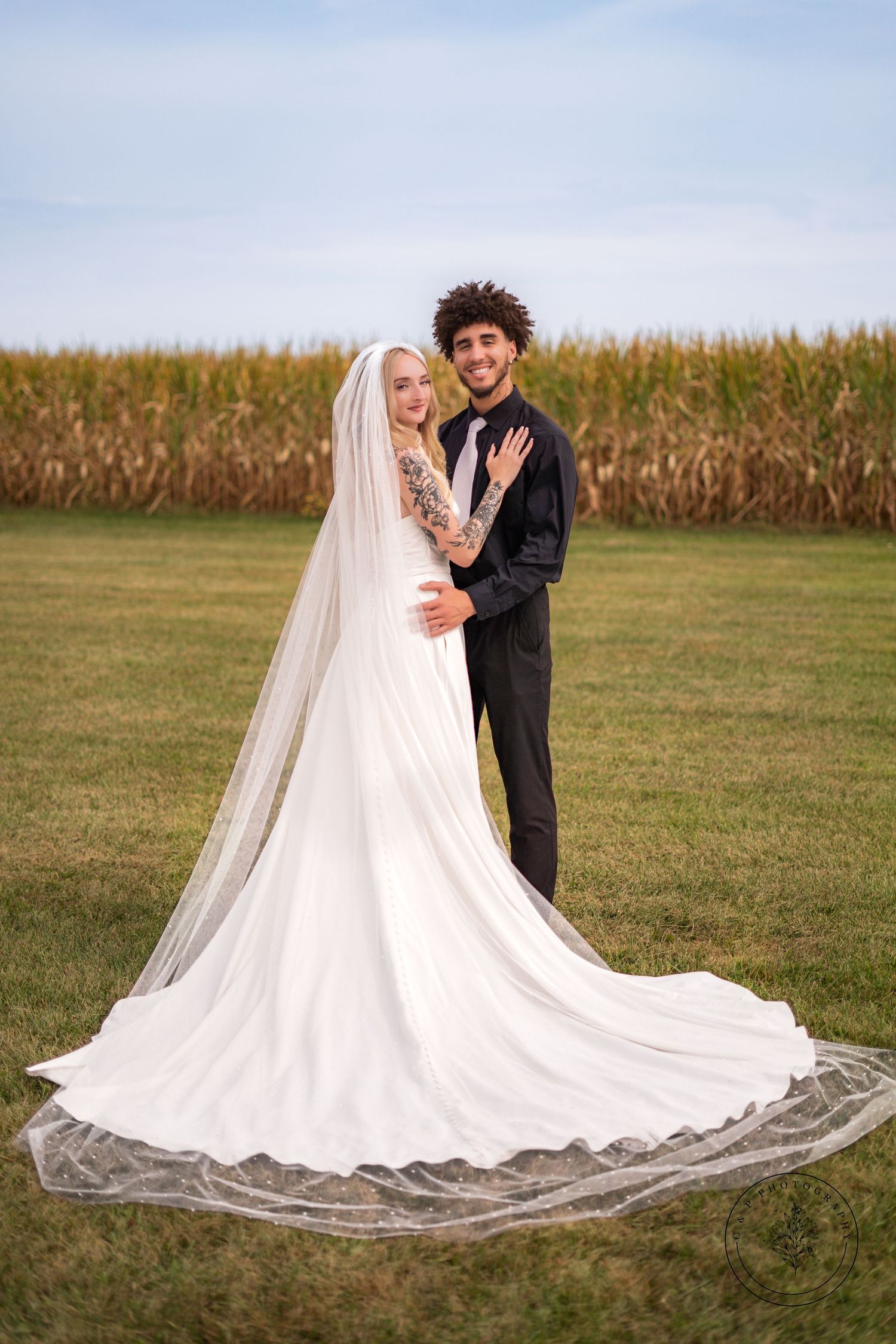 Bride in flowing white gown with long veil poses with groom near cornfield.