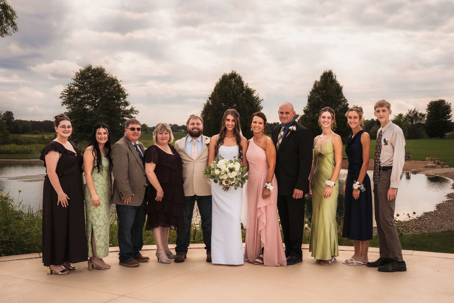 Wedding party poses outdoors near a pond, wearing formal attire including gowns and suits, under a cloudy sky.