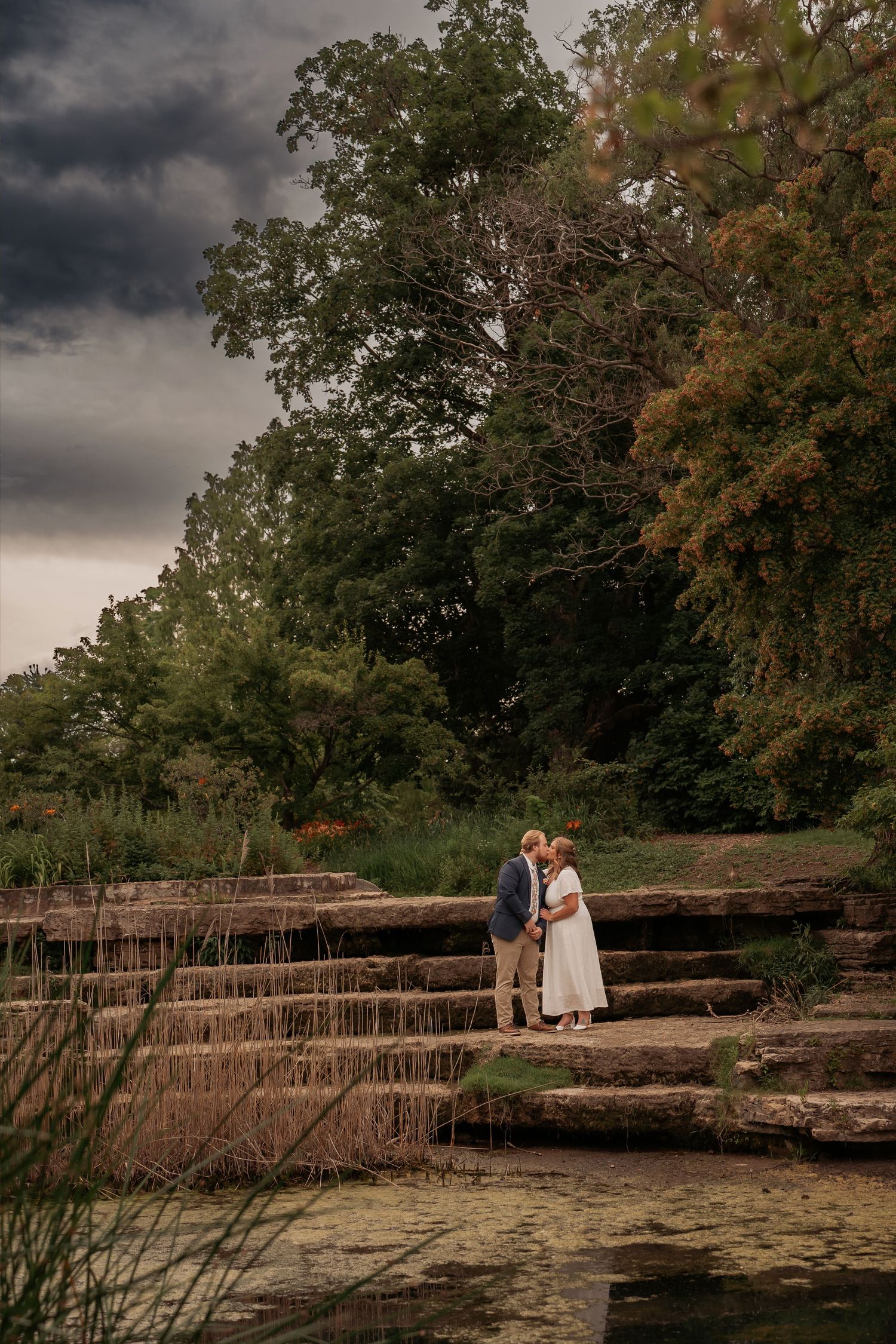 Couple shares a romantic moment on stone steps surrounded by lush greenery under dramatic stormy skies.