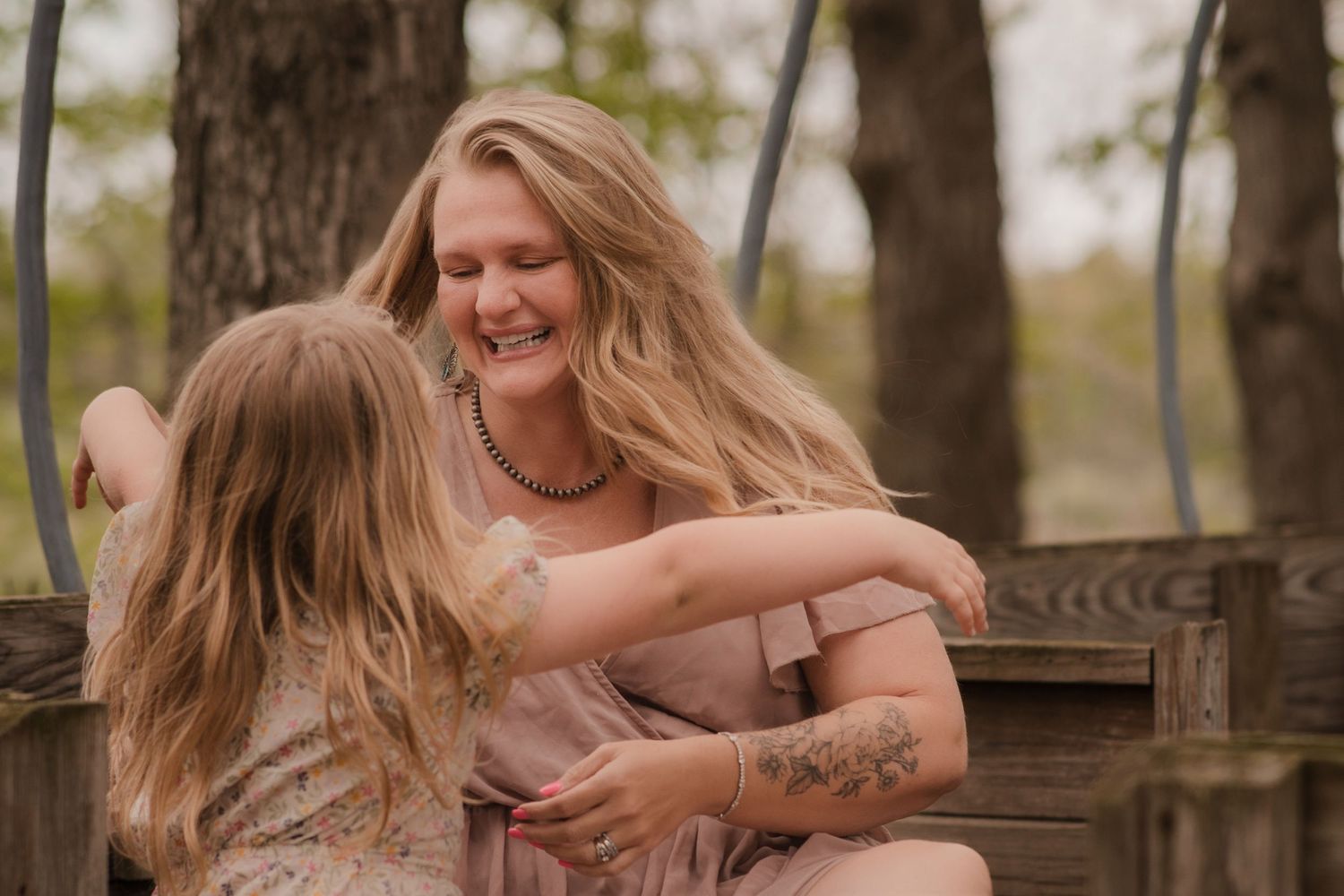 A laughing woman with tattoos hugs a young girl on a wooden swing outdoors among trees.