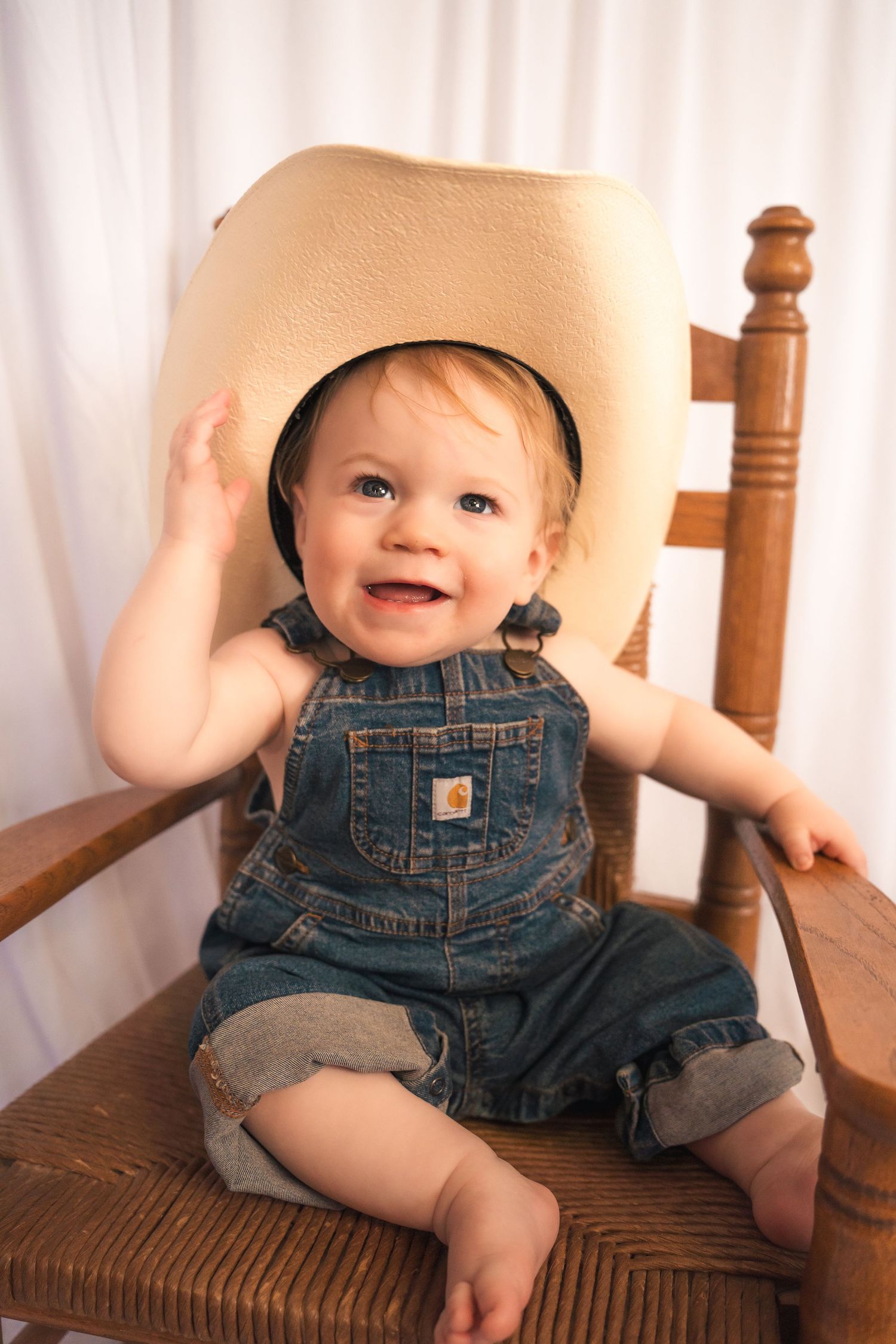 Baby in denim overalls sits and smiles on wooden chair against white backdrop.