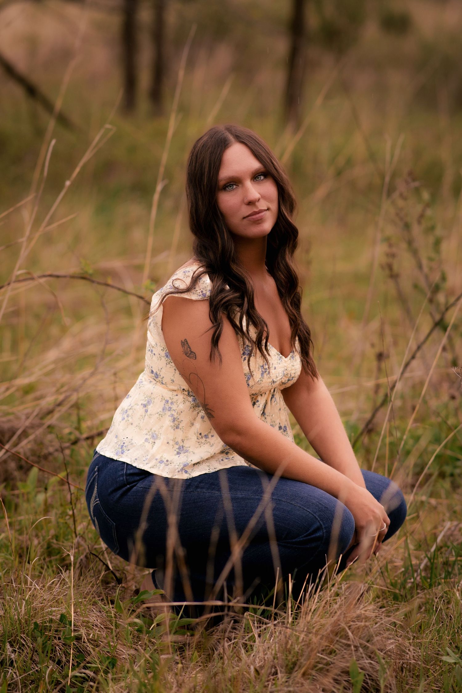 A young woman with long dark hair sits crouched in a field of tall dry grass, wearing a floral top and blue jeans.