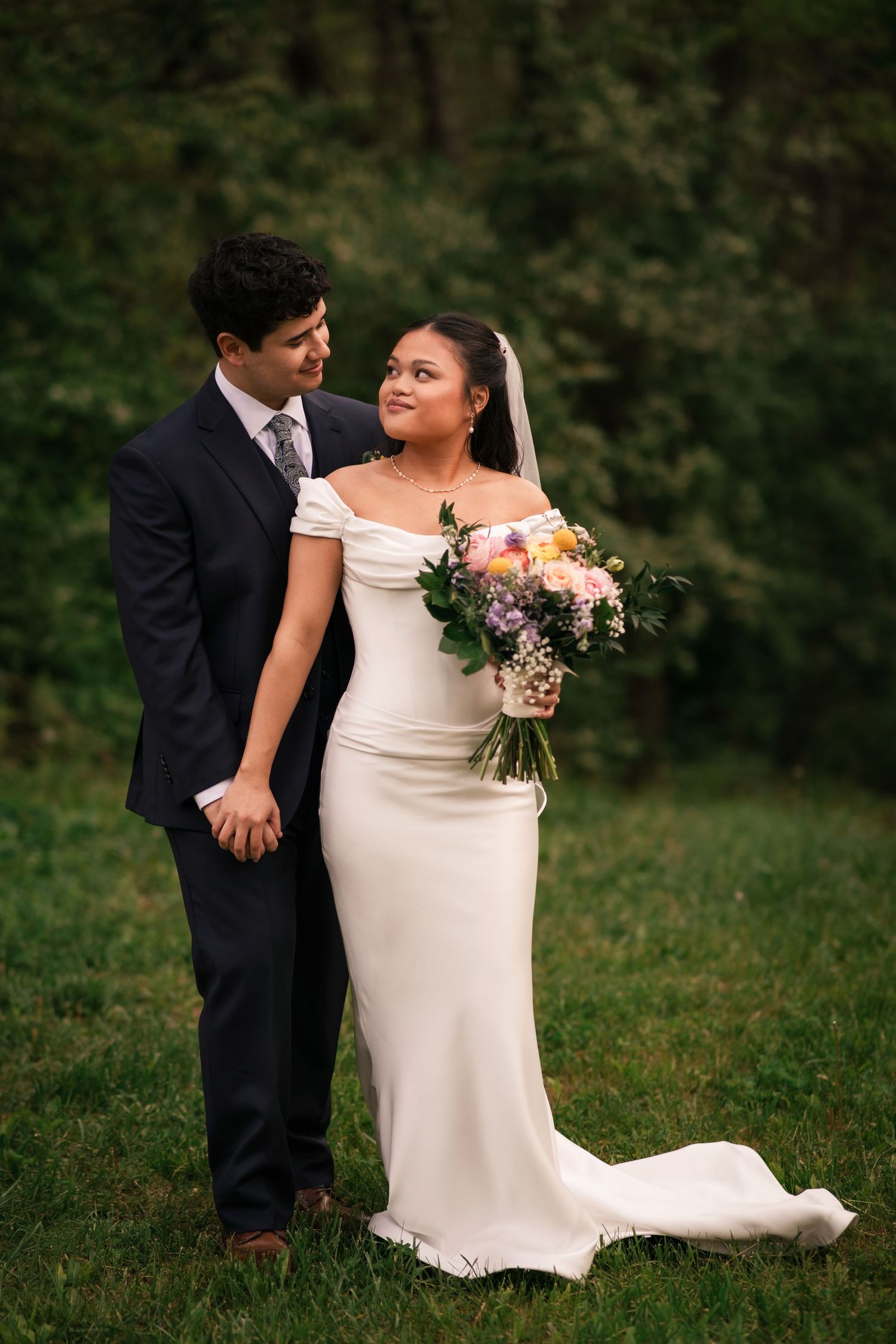 A bride and groom pose together on green grass, she holds a colorful bouquet in a white off-shoulder gown.