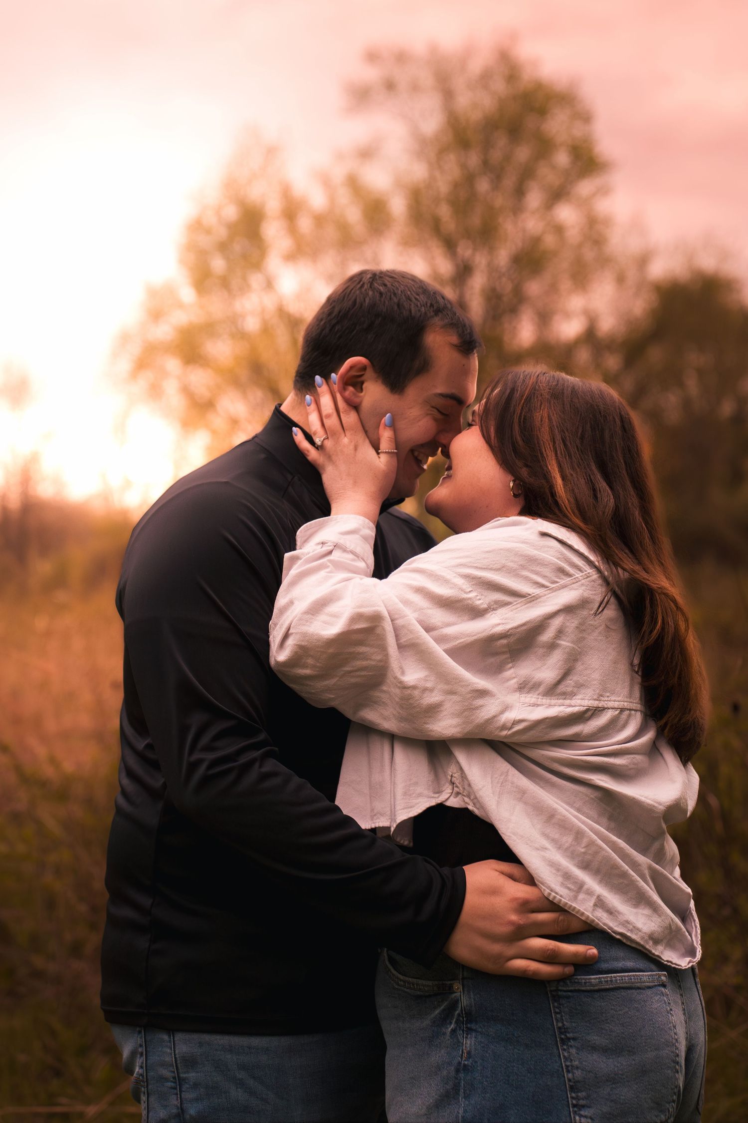 Couple sharing a romantic moment at sunset in a field, woman in white shirt embracing man in black jacket.
