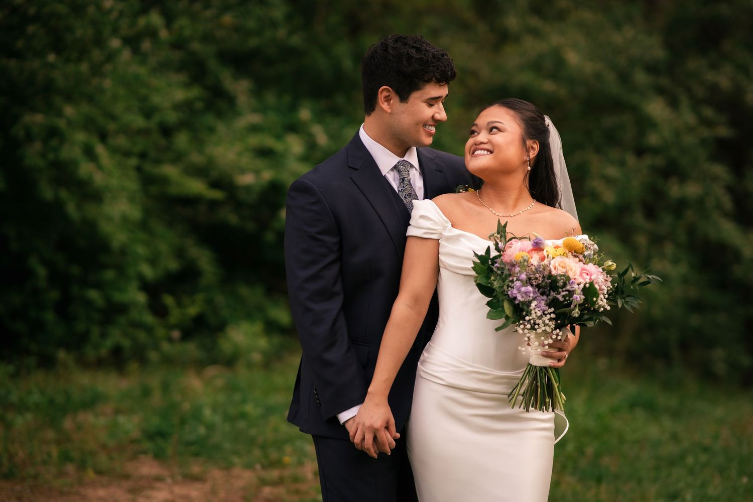Bride in white gown holding colorful bouquet smiles at groom in dark suit, standing outdoors with lush greenery backdrop.