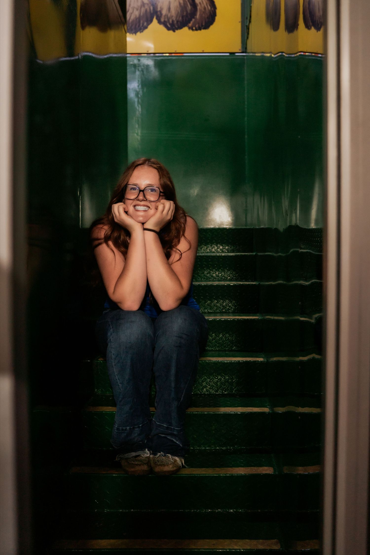 A smiling teenage girl with glasses sits on green stairs, resting her chin on her hands in a cozy stairwell.