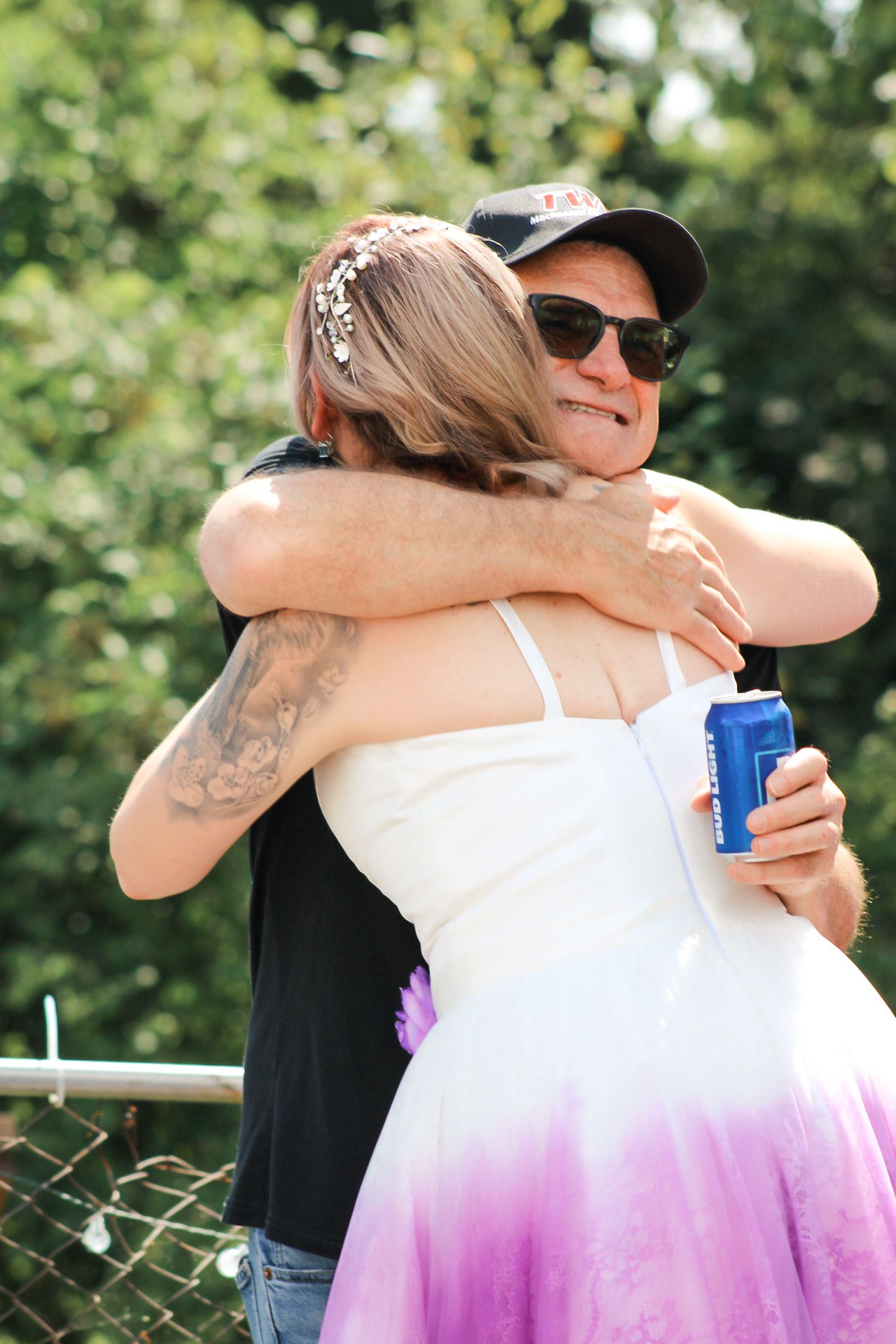Two people share a warm embrace at an outdoor event while one holds a blue drink can.