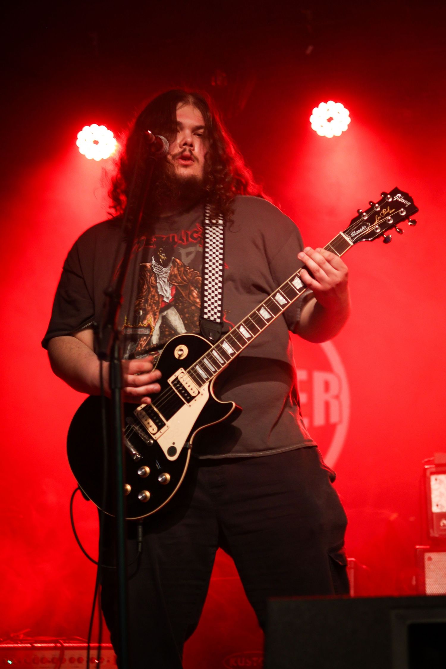 A long-haired musician playing a black Les Paul guitar on stage with dramatic red lighting.