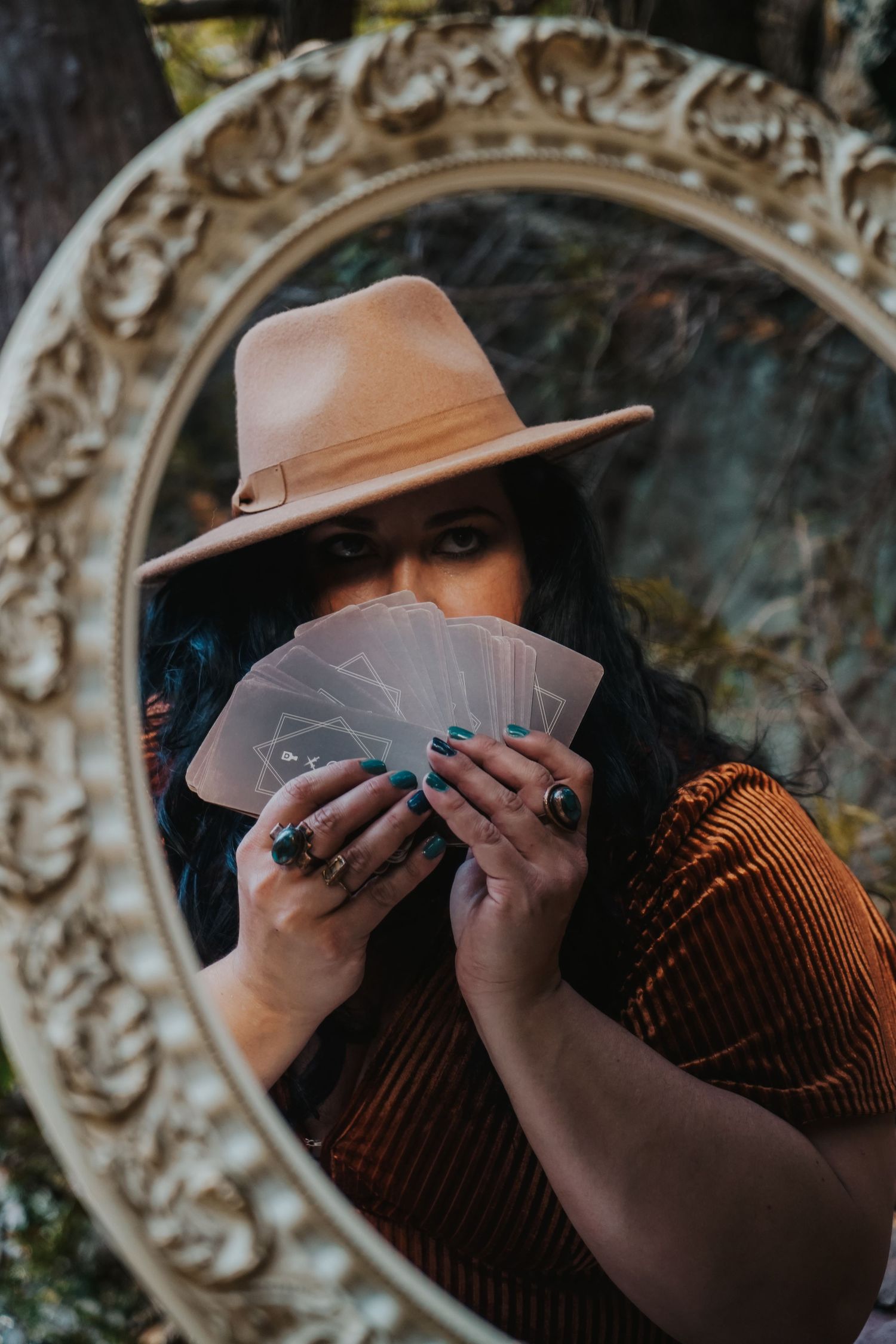 Mysterious figure in tan fedora holds playing cards in front of ornate vintage mirror in atmospheric setting.