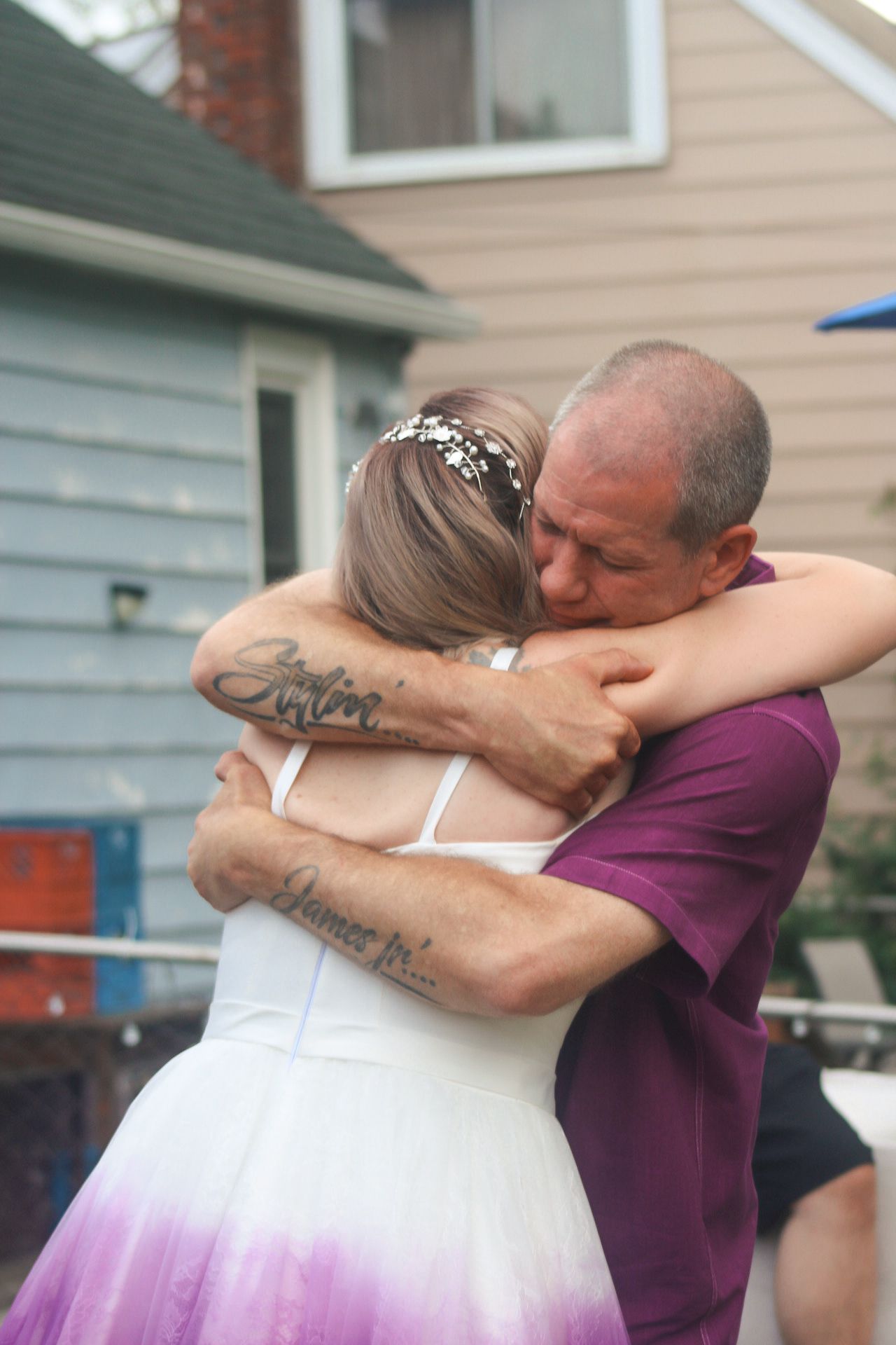 Two people share an emotional embrace outside a house, one wearing a white and purple gradient wedding dress with a hair accessory.
