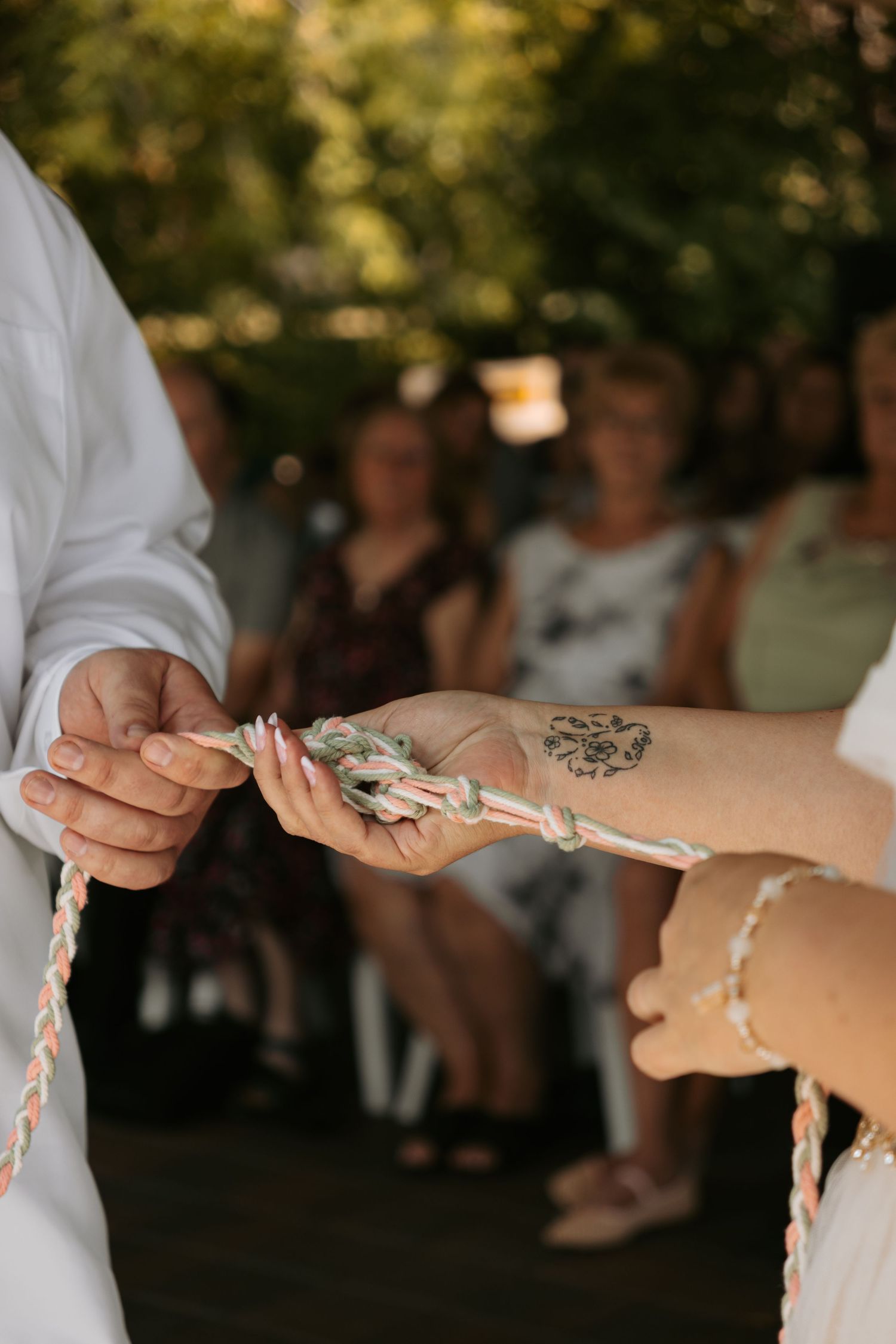 Close-up photo of hands tying a decorative rope knot during a wedding ceremony in soft outdoor lighting.