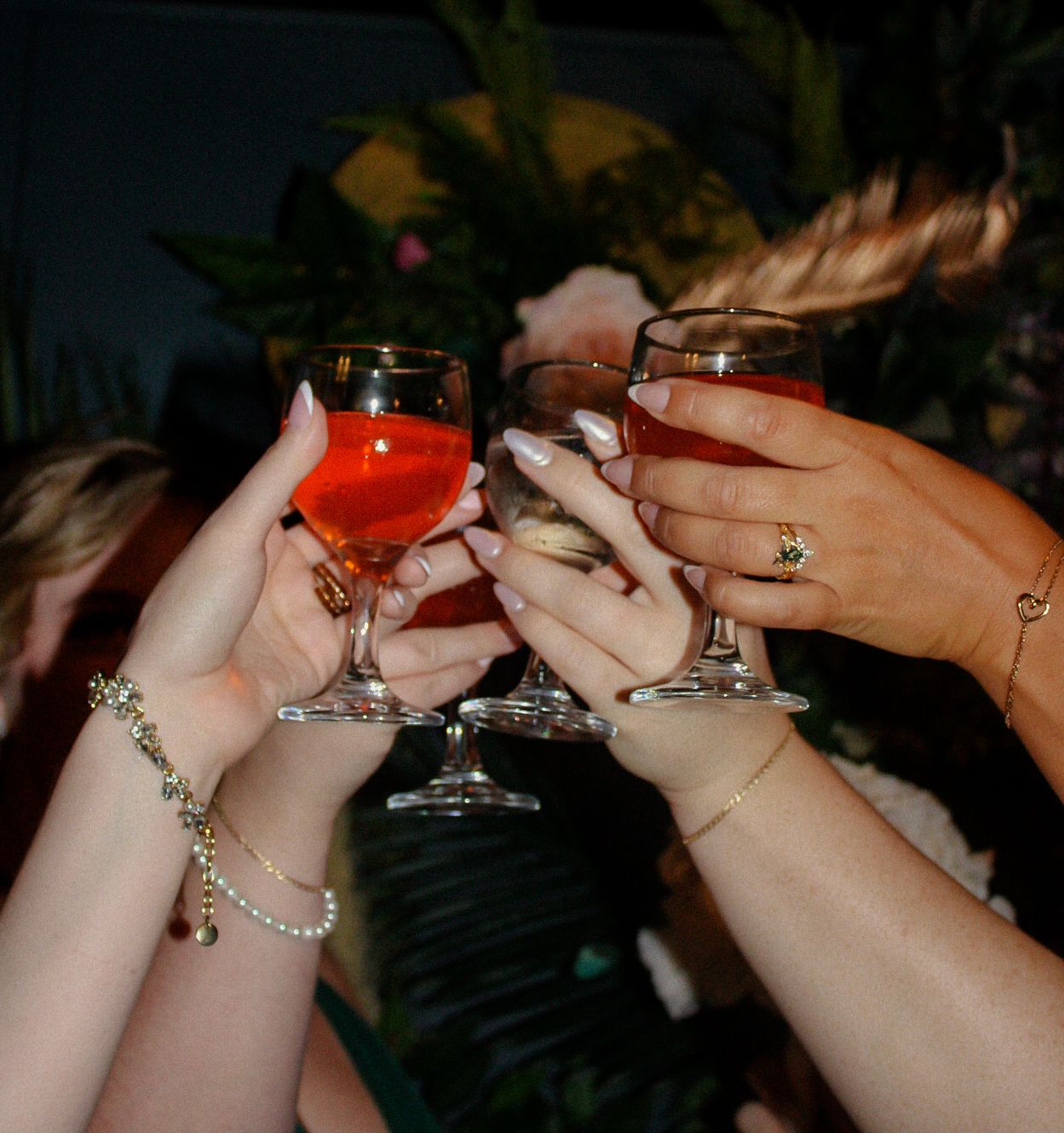 Multiple hands raise cocktail glasses in a cheers toast, with jewelry and bracelets visible in a dark setting.