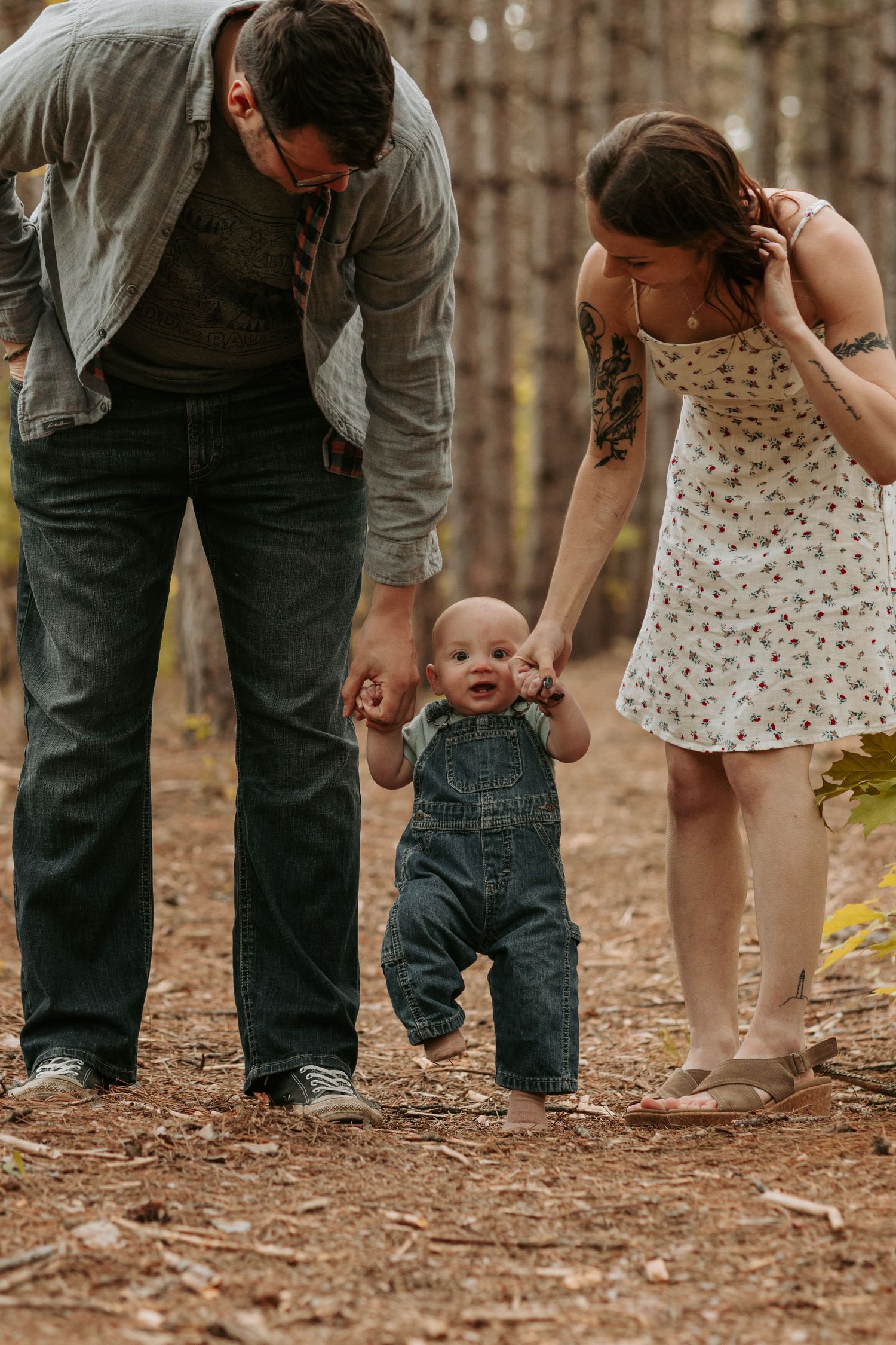 Family assists baby taking first steps in overalls while walking through a forest of pine trees on autumn day.