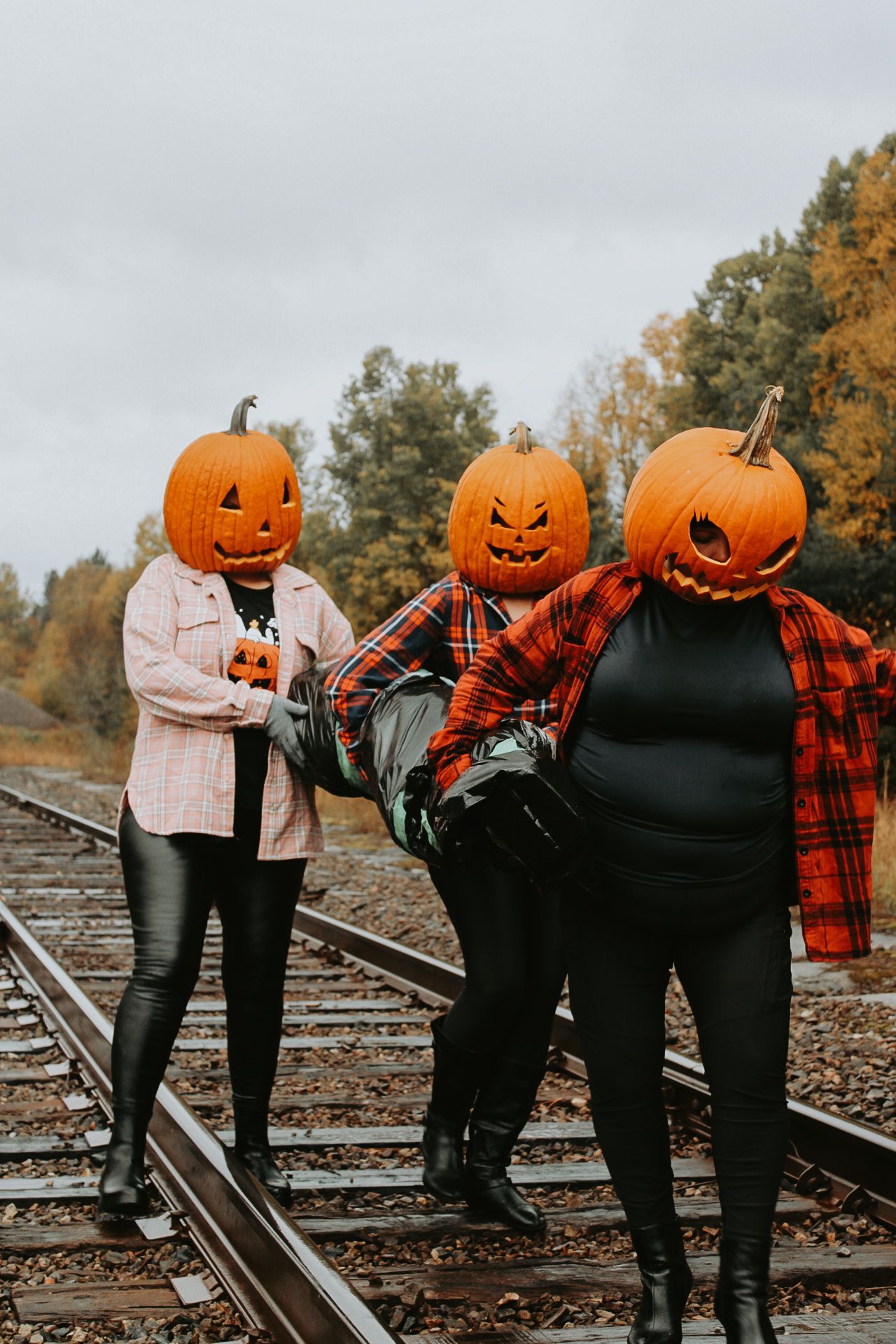 Three friends in pumpkin head costumes posing on train tracks during autumn.