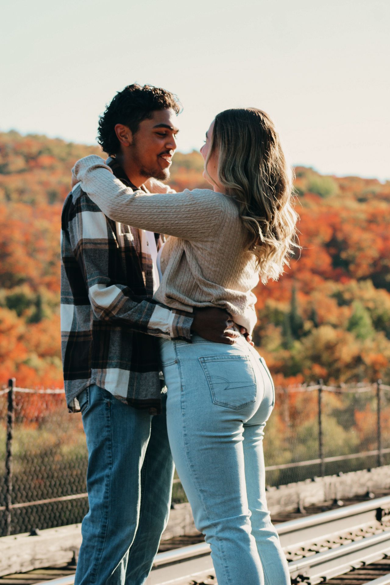 Romantic couple embracing on bridge with vibrant fall foliage in background.