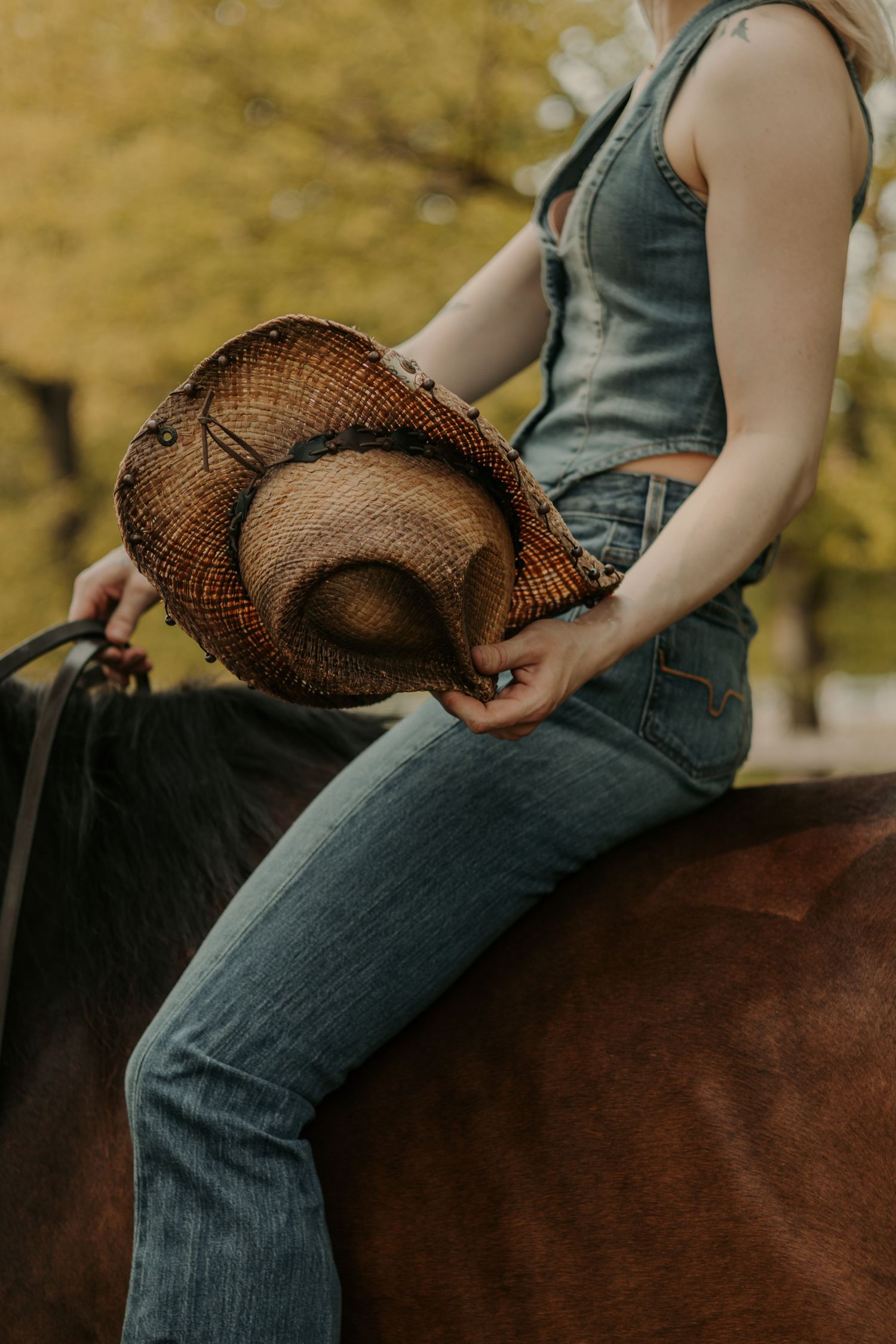 Person in denim overalls riding horseback while holding a brown straw cowboy hat against autumn backdrop.