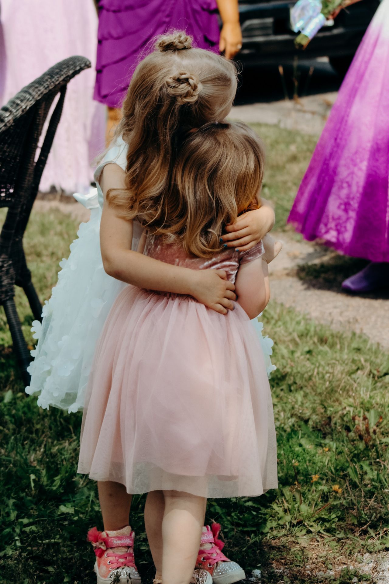 Two young children in pastel dresses share a sweet hug at what appears to be a formal outdoor event.