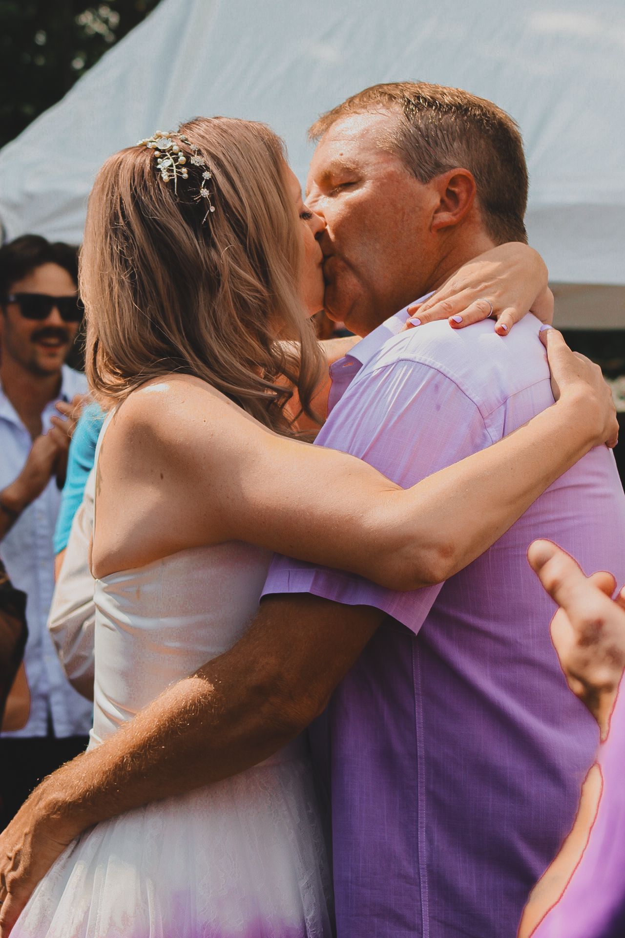 A couple shares their first kiss as newlyweds under a canopy at an outdoor wedding celebration.
