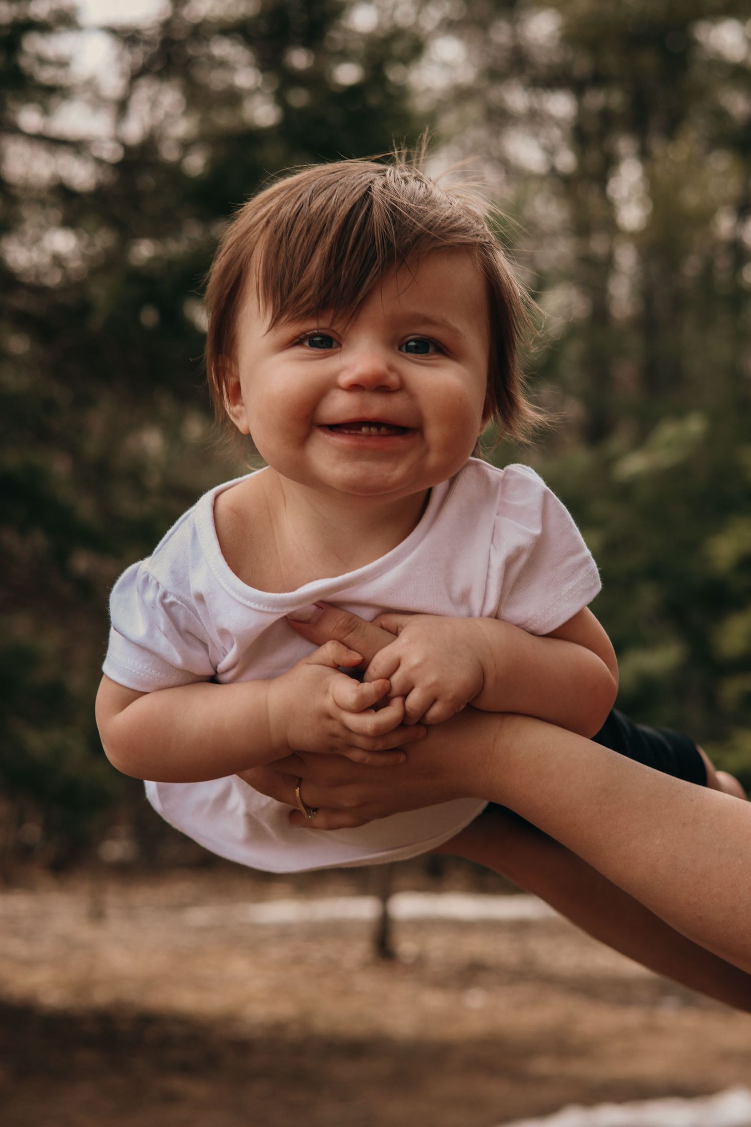 A baby wearing a white top smiles joyfully while being held up in the air outdoors with trees in the background.