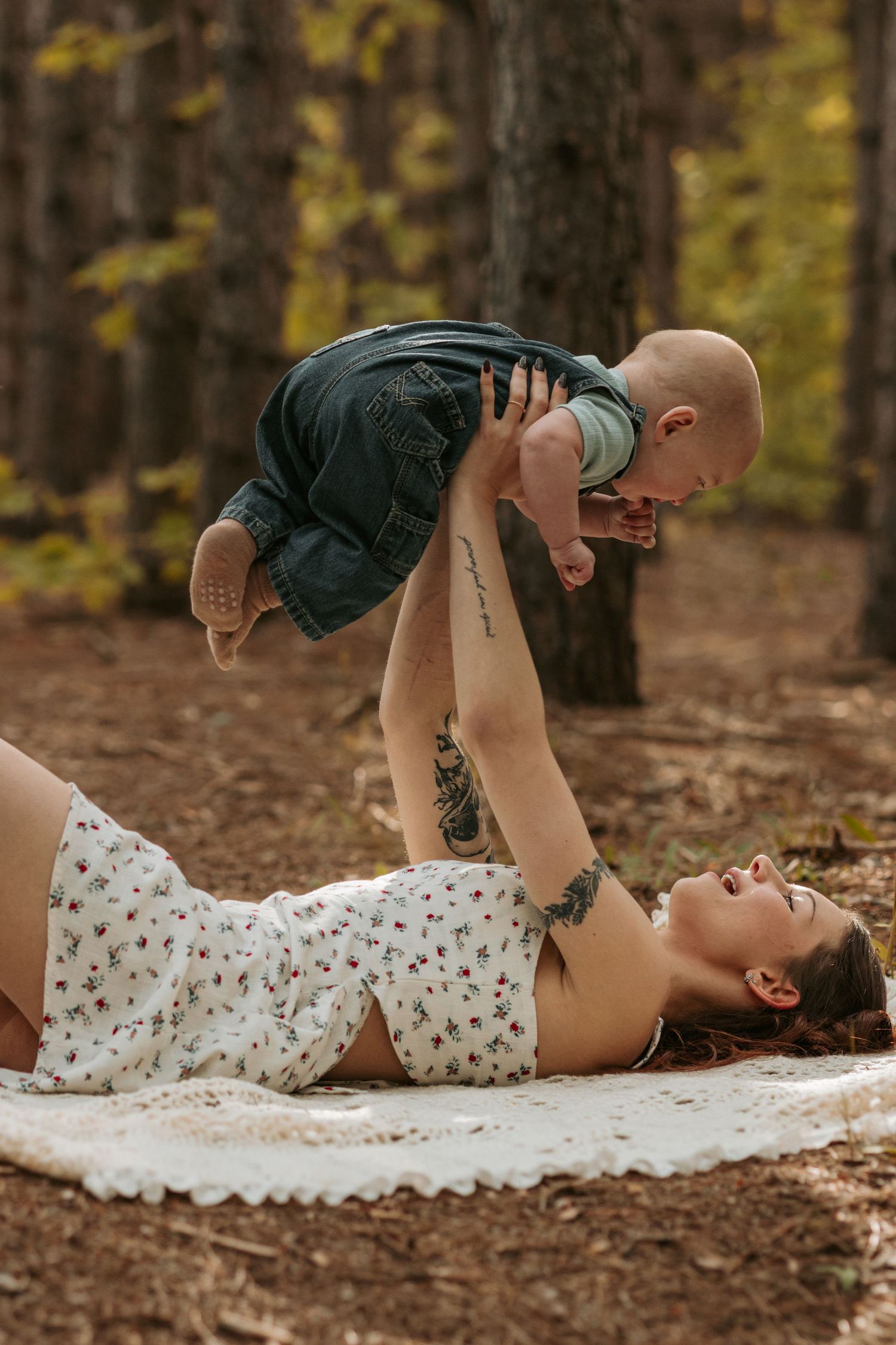 A parent and child share a playful moment while lying on a blanket in an autumn forest setting.