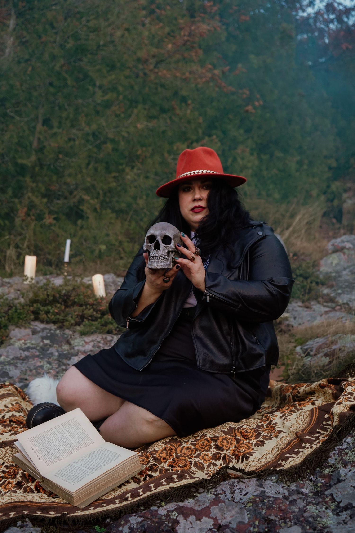 A person in black clothing and red hat sits on a blanket with a book and decorative skull in an autumn forest setting.