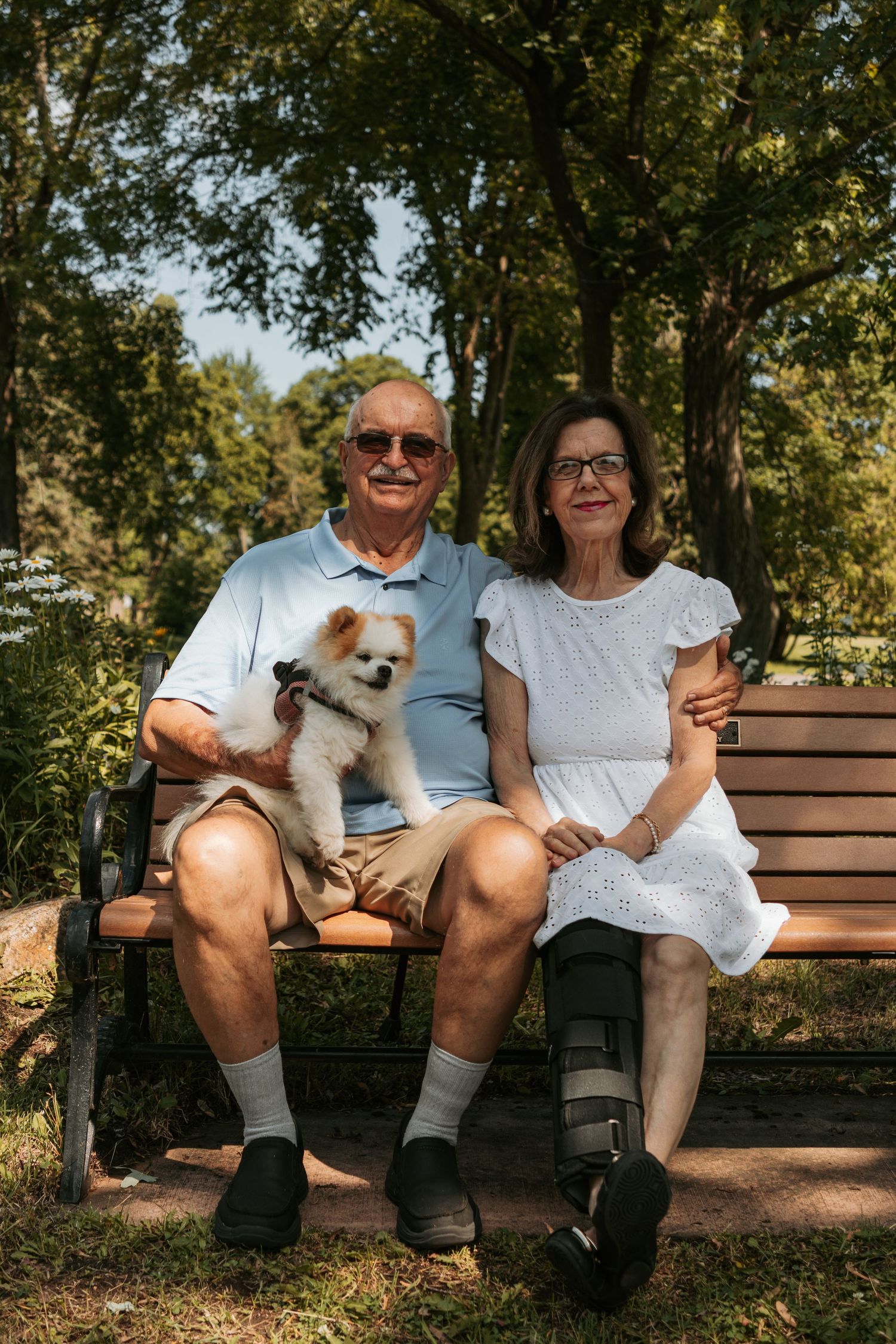 People sitting on a wooden park bench together on a sunny day surrounded by trees and dappled sunlight.