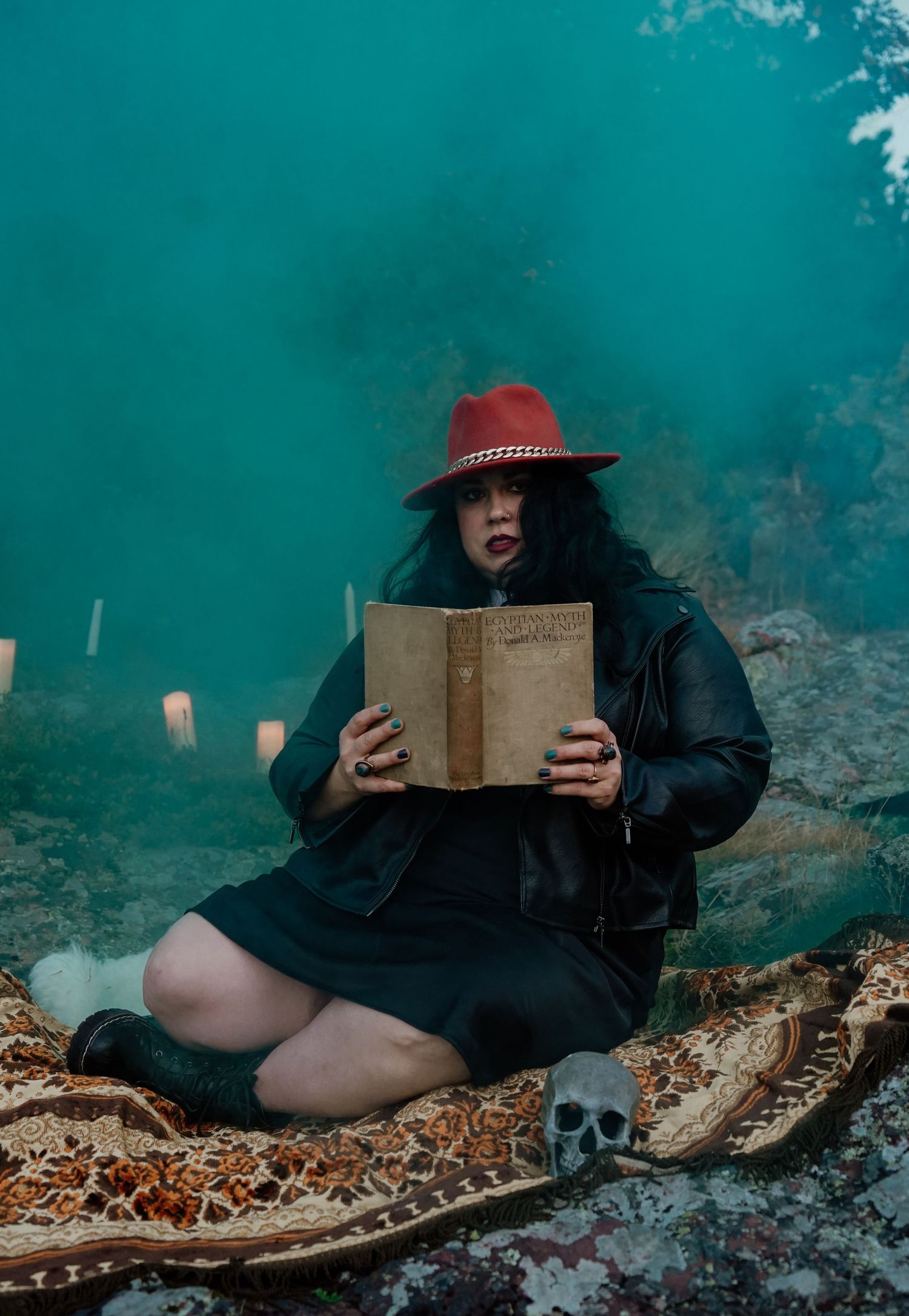 Figure in red hat reads old book while sitting on ground with candles and skull in moody teal setting.