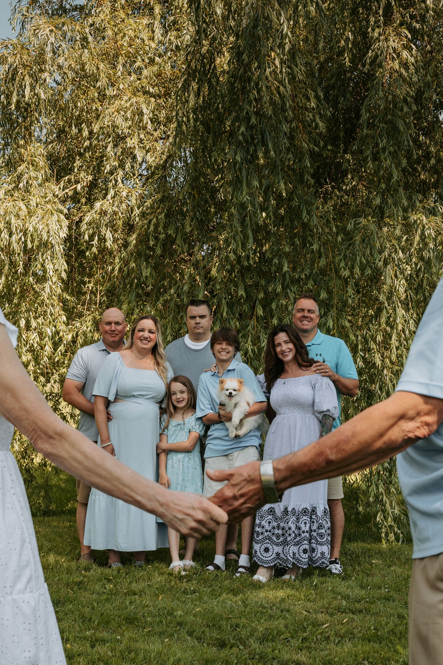 Extended family wearing coordinated light blue outfits poses together under a weeping willow tree during outdoor photo session.
