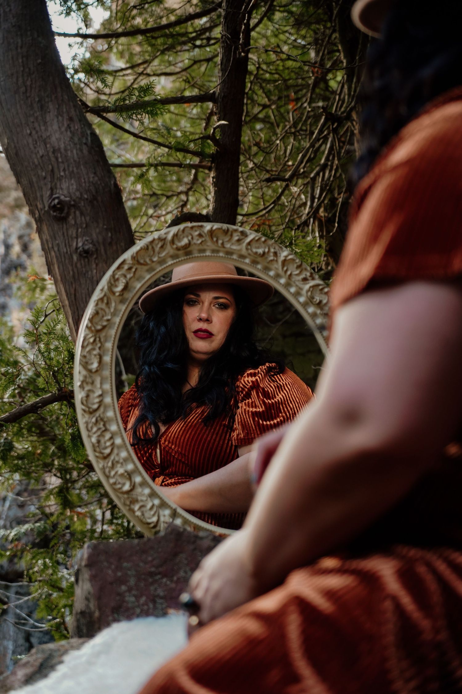 Ornate vintage mirror reflecting a scene of a woman in a red dress and wide-brimmed hat among trees in moody lighting.
