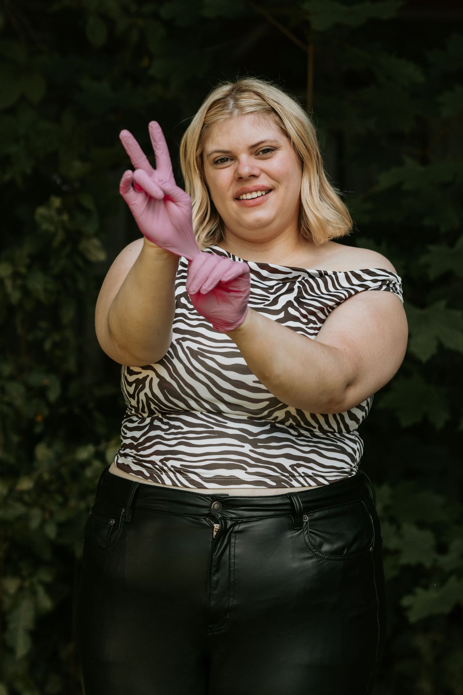 Person wearing a zebra print top and black pants gives peace sign while posing outdoors against green foliage.