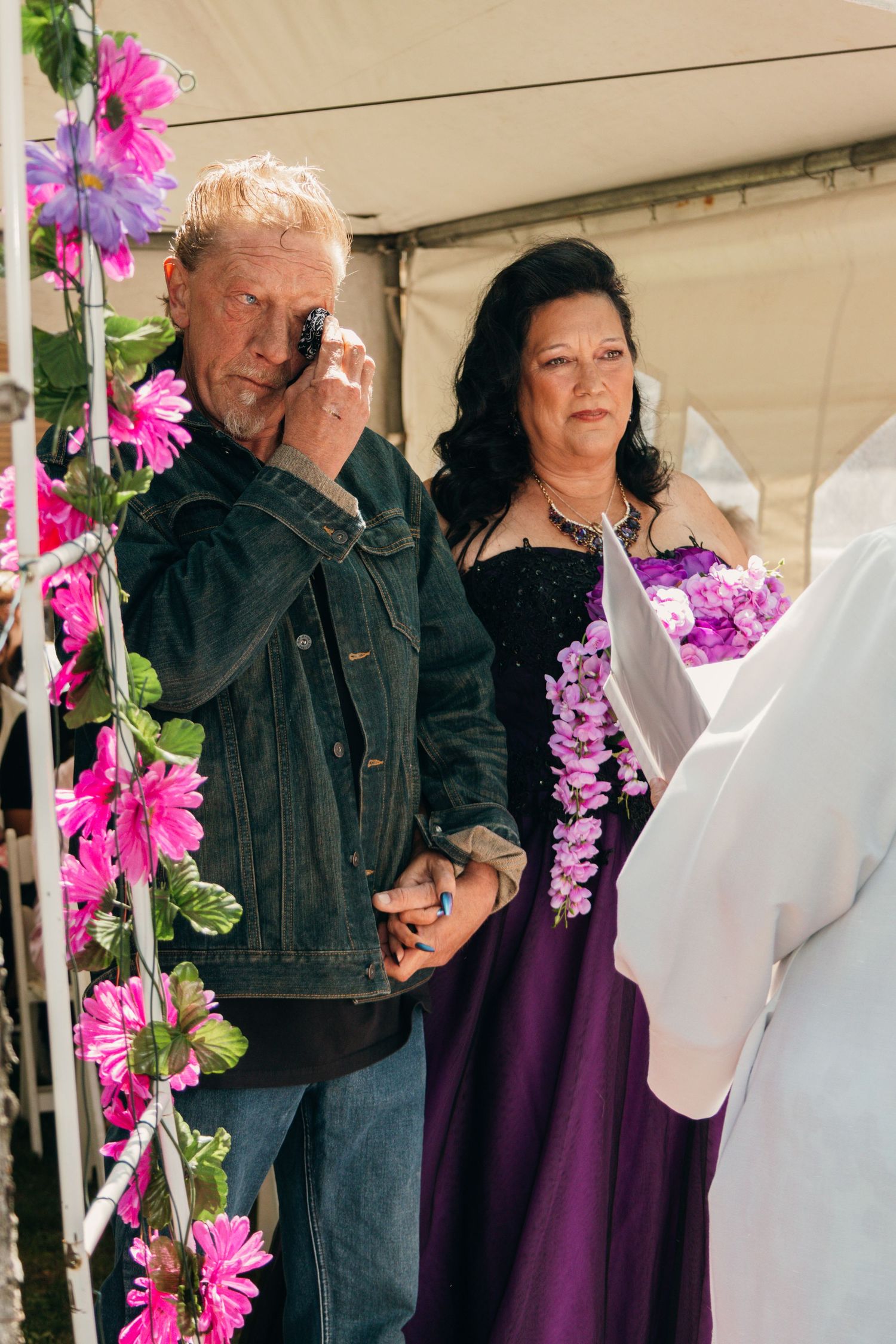 Wedding couple stands at altar under purple floral arch during outdoor ceremony.