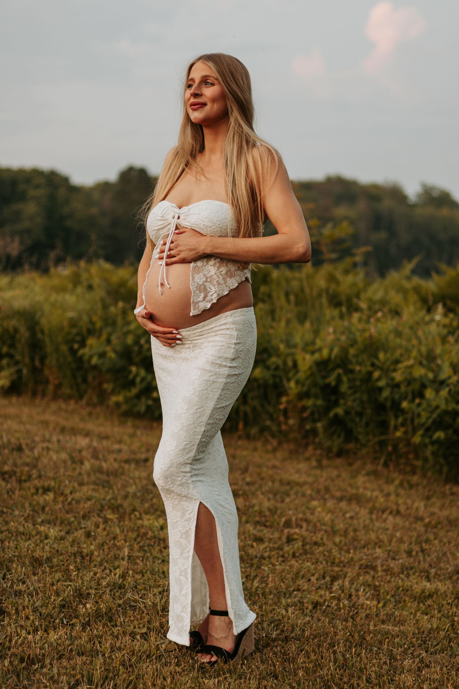 Maternity photo of expectant mother in white dress posing in golden field at sunset.