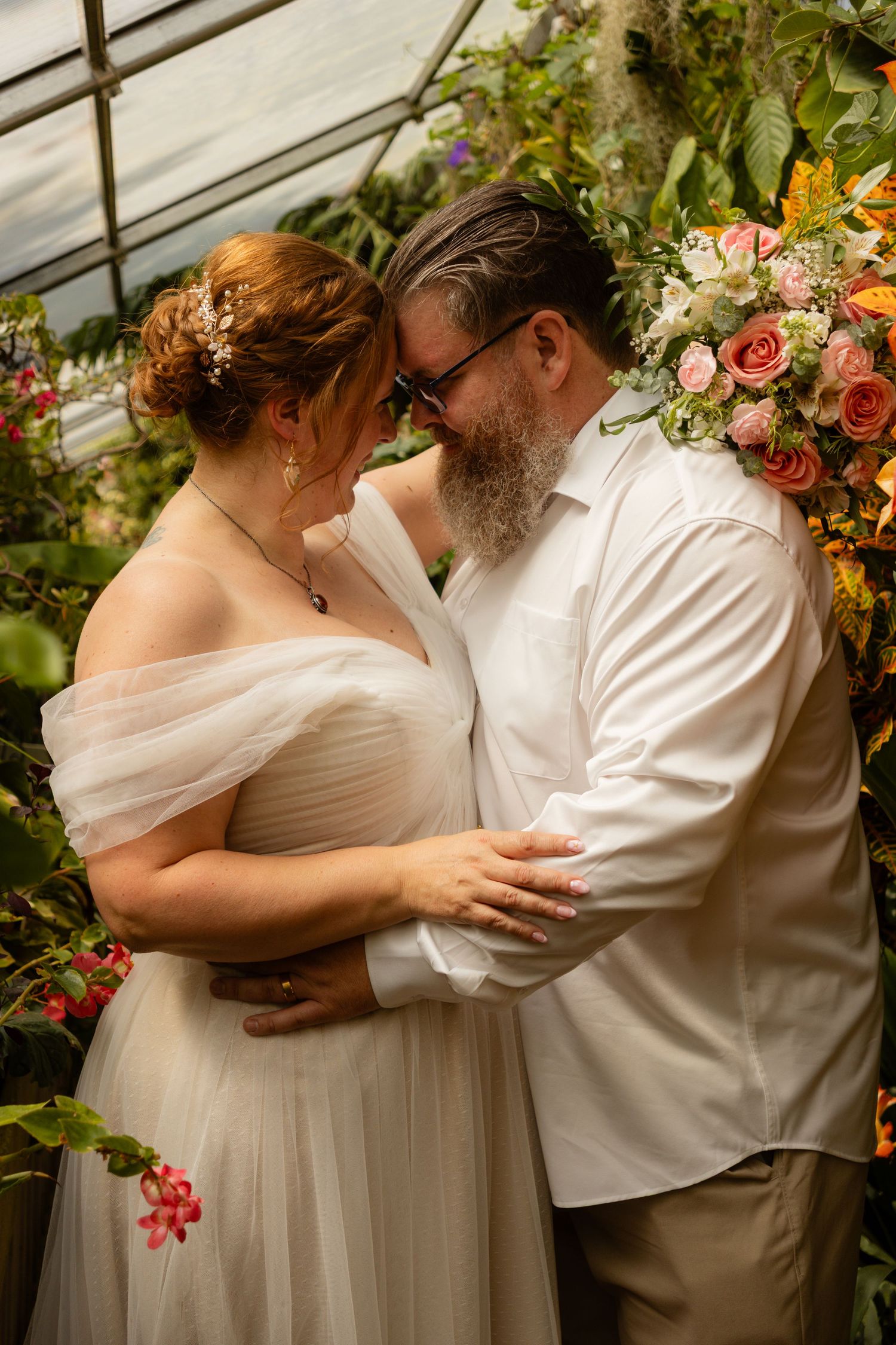 A romantic wedding couple embraces in a greenhouse surrounded by lush floral arrangements and greenery.