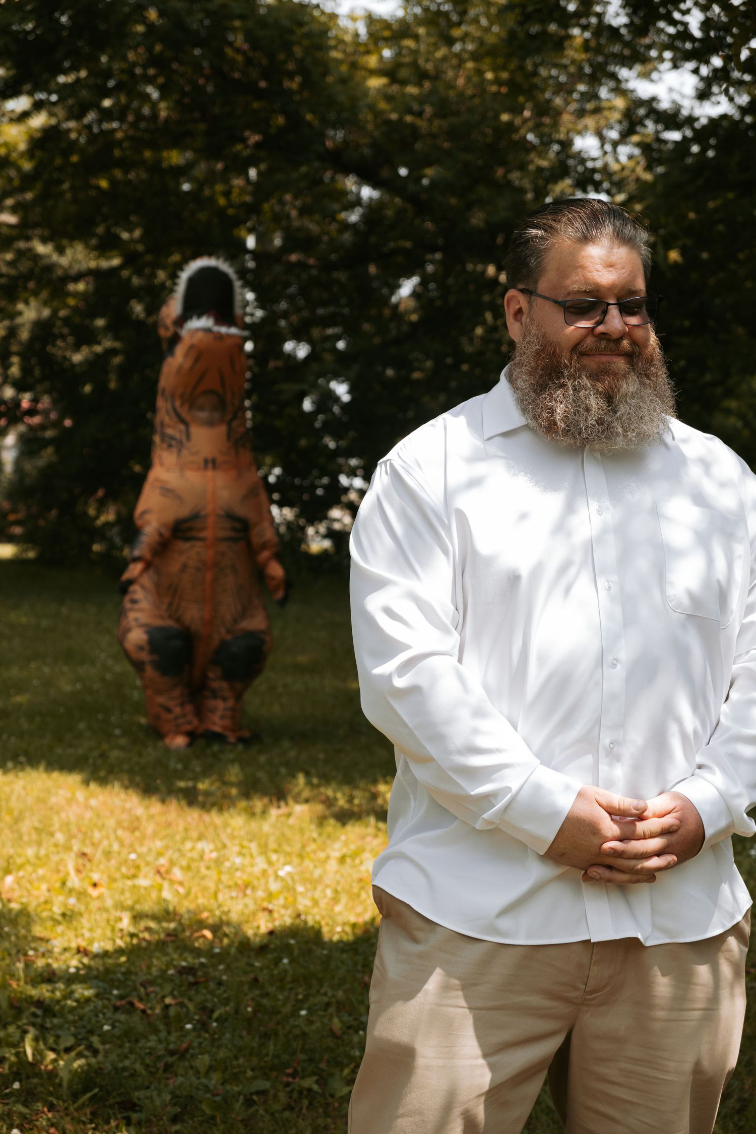 A person in white shirt and sunglasses stands while a T-Rex dinosaur costume appears to jump in the background outdoors.