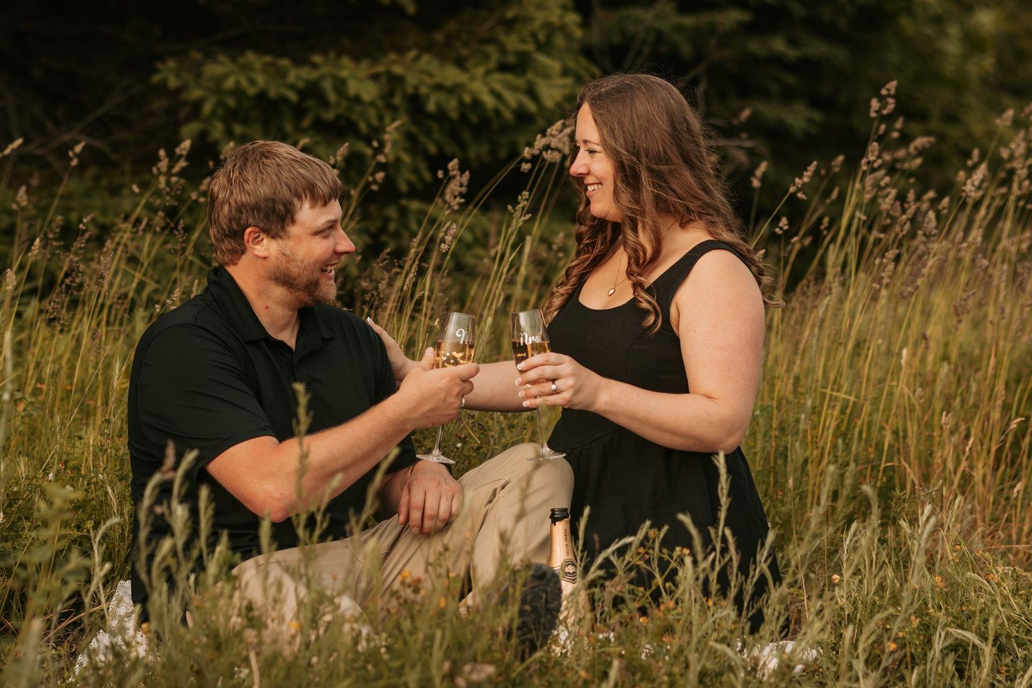 A couple shares a romantic moment kneeling in a golden meadow at sunset during what appears to be a proposal.