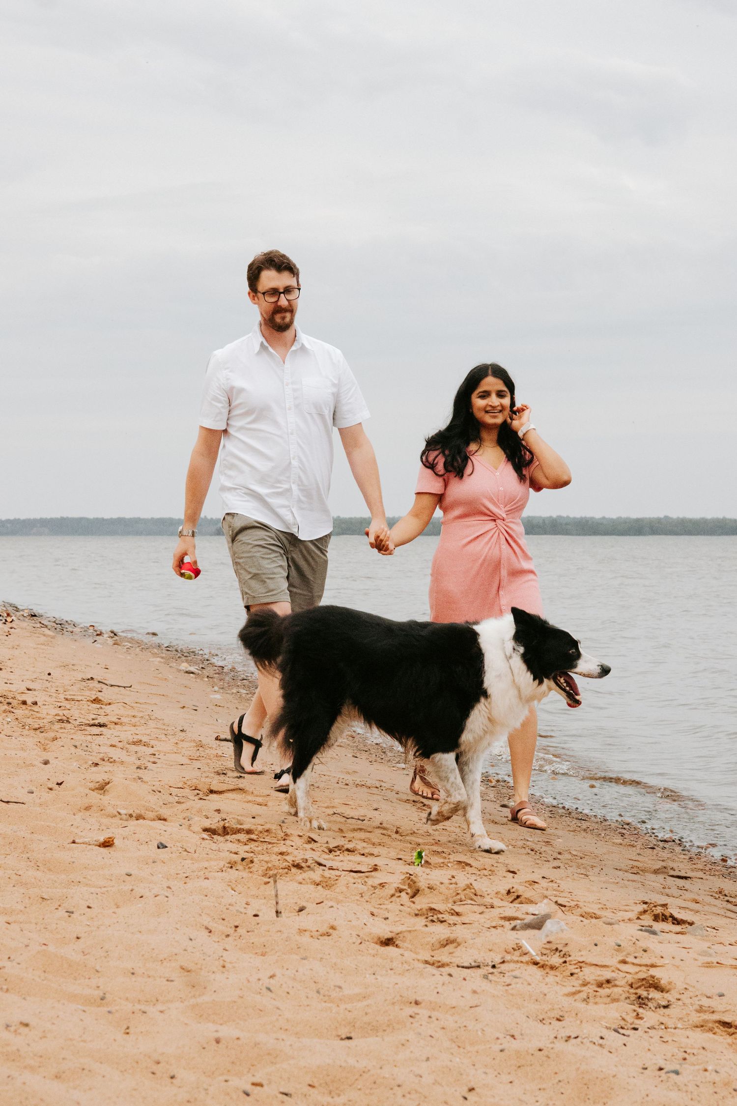 A couple walks along a sandy beach with their Border Collie dog as the waves lap at the shore.