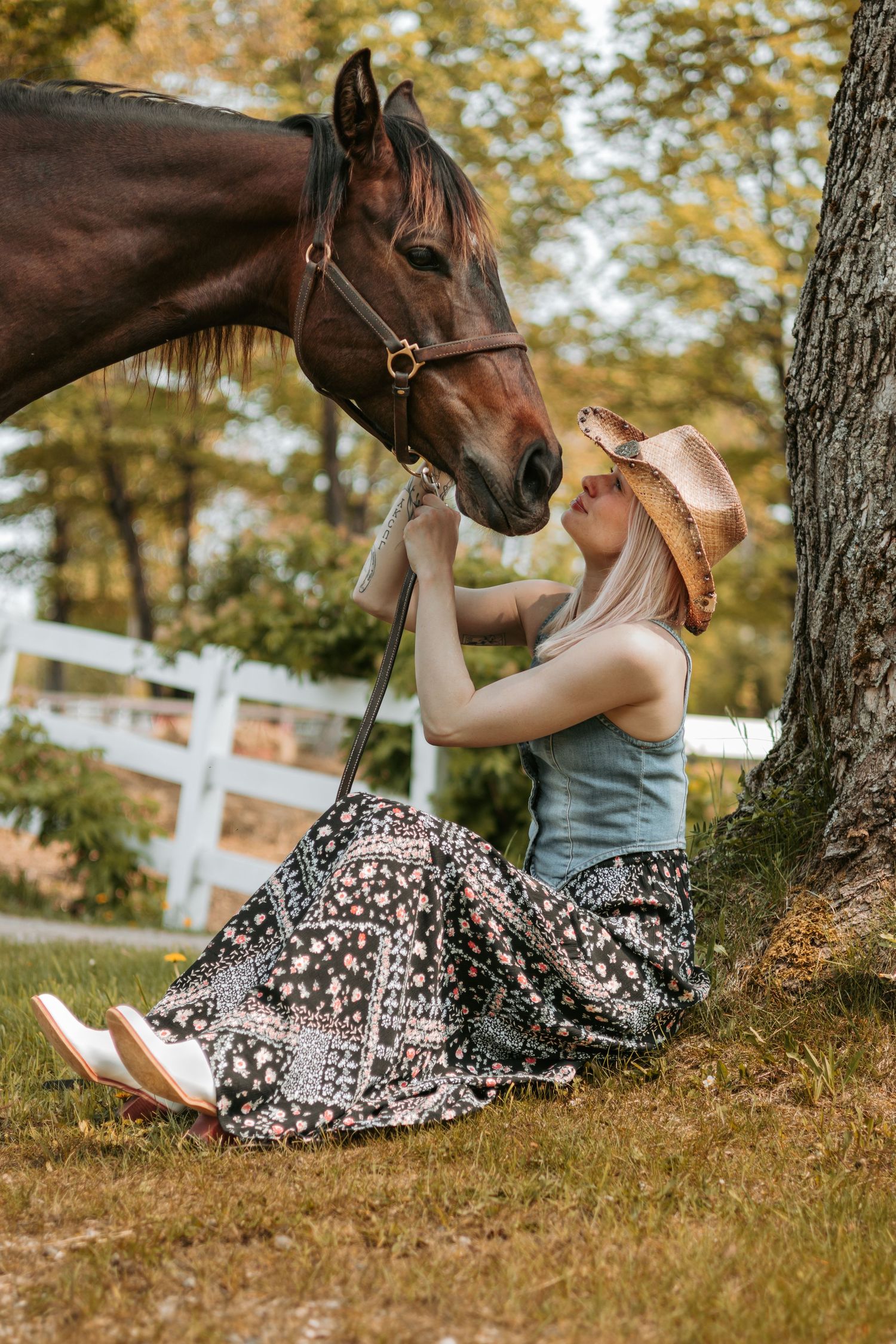 A brown horse nuzzles close to someone sitting in a floral skirt and cowboy hat by a white fence on a sunny day.