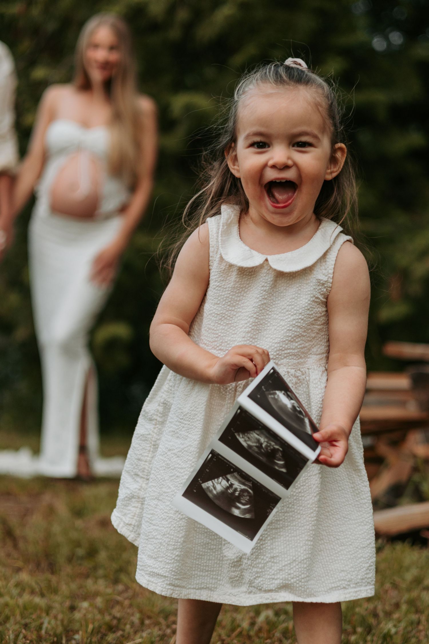 Excited child in white dress holds ultrasound photos while pregnant figure stands in background.