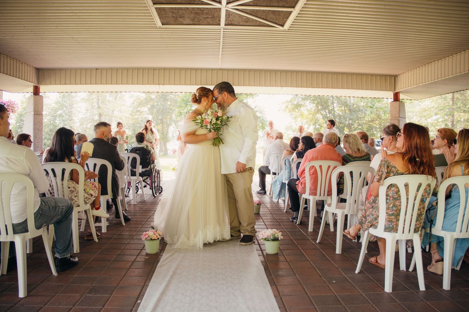 A romantic outdoor wedding ceremony under a covered pavilion with white chairs and natural lighting.