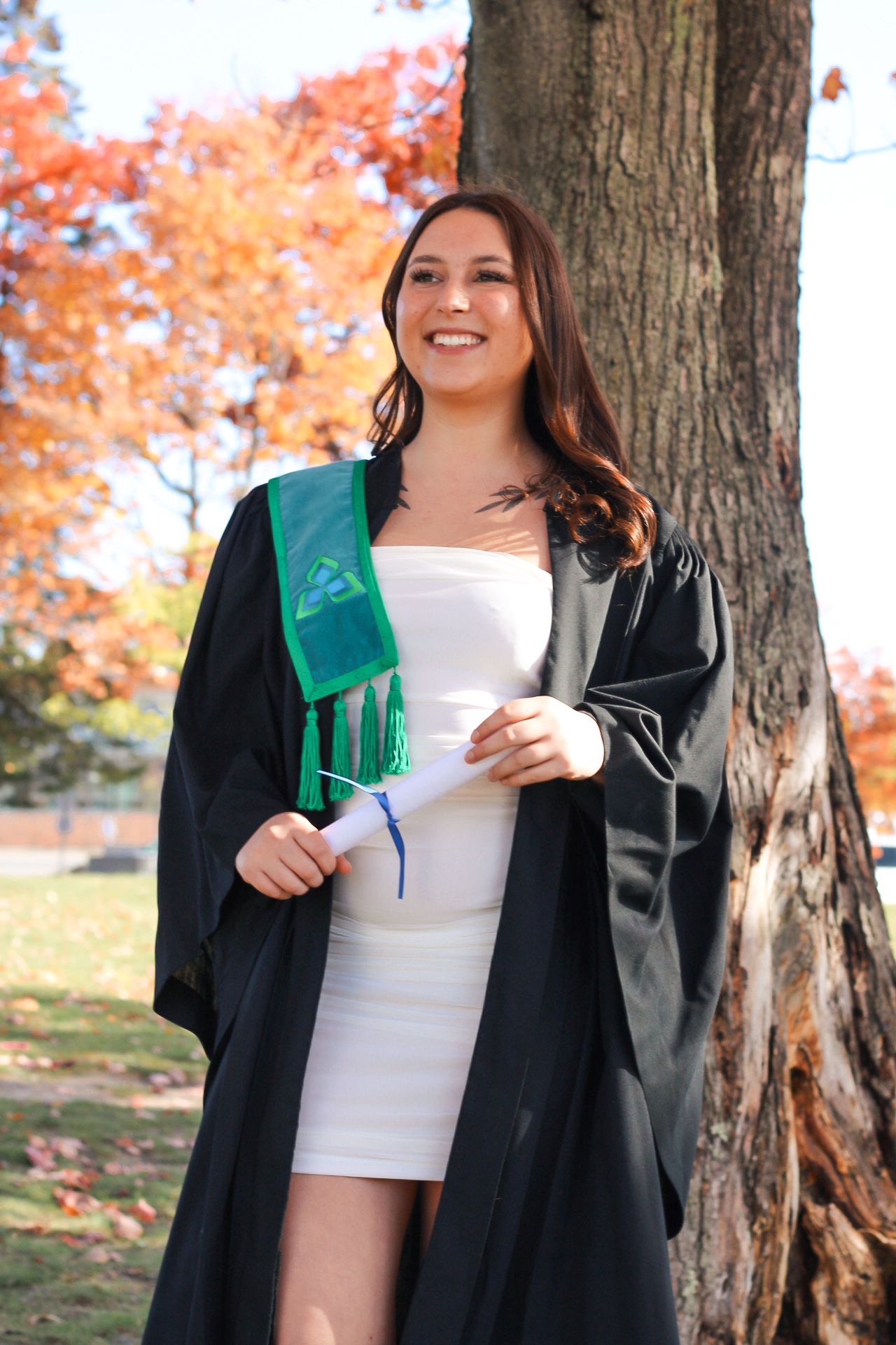 Graduate in white dress holding diploma and green scarf poses by tree with autumn leaves.